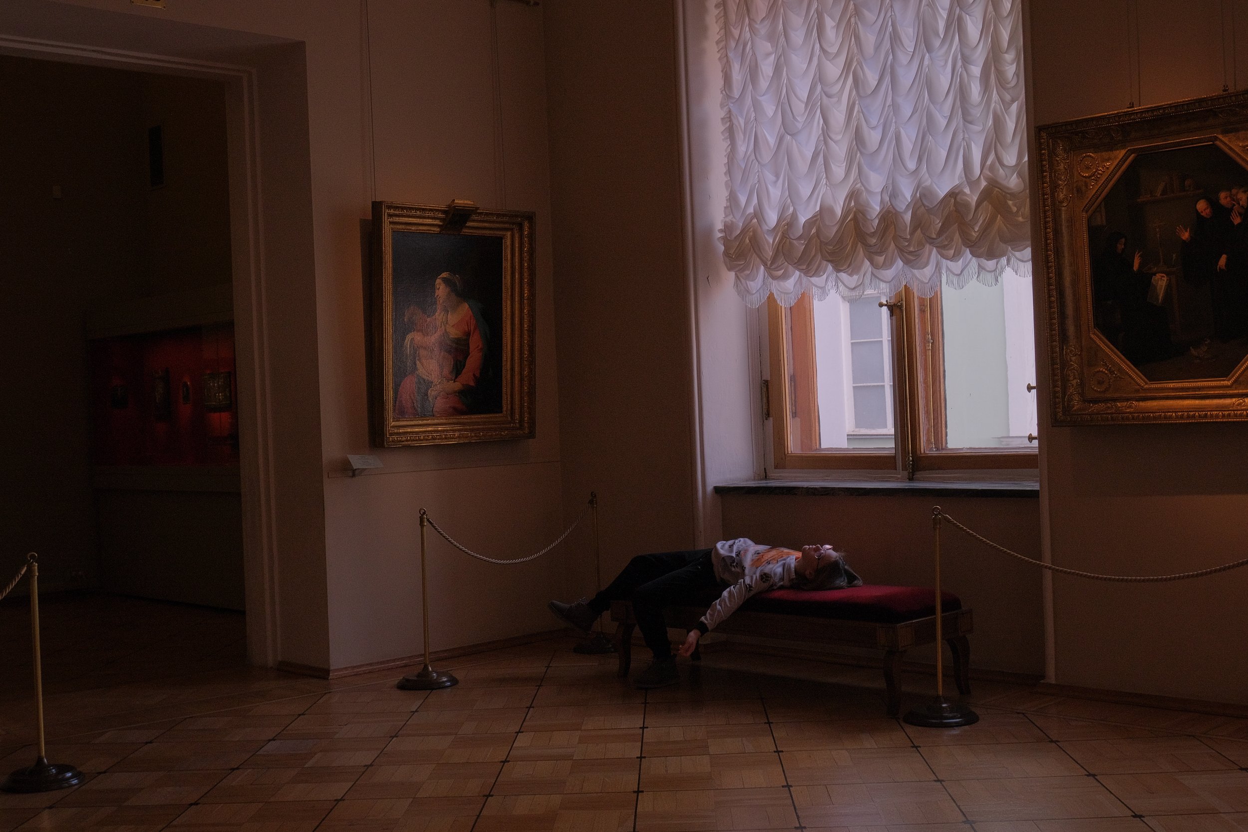 Person lying on a bench in an art gallery, surrounded by framed paintings on the walls, with natural light coming through a window.
