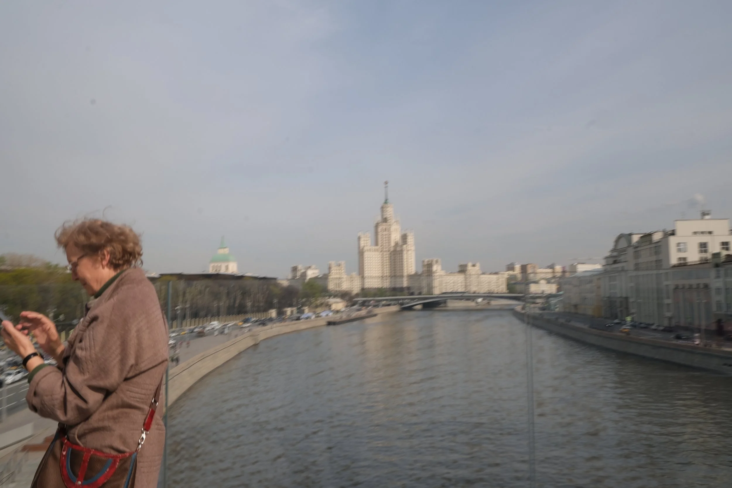 A woman with curly hair wearing a brown coat, standing by a river, using her phone, with a city skyline and bridge in the background.