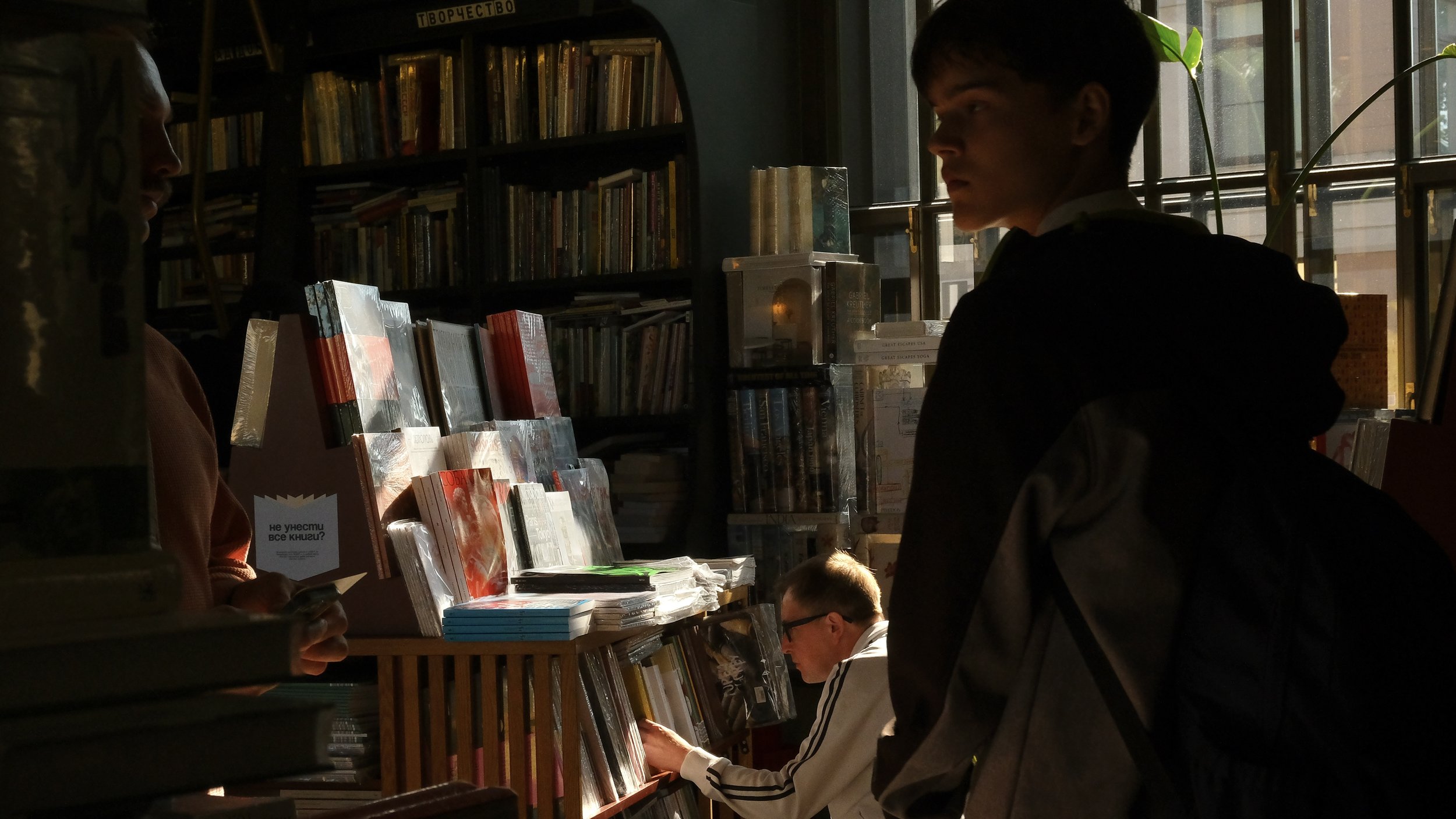 Three people inside a bookstore,  with dark shadows cast on their faces, surrounded by books on shelves and tables.
