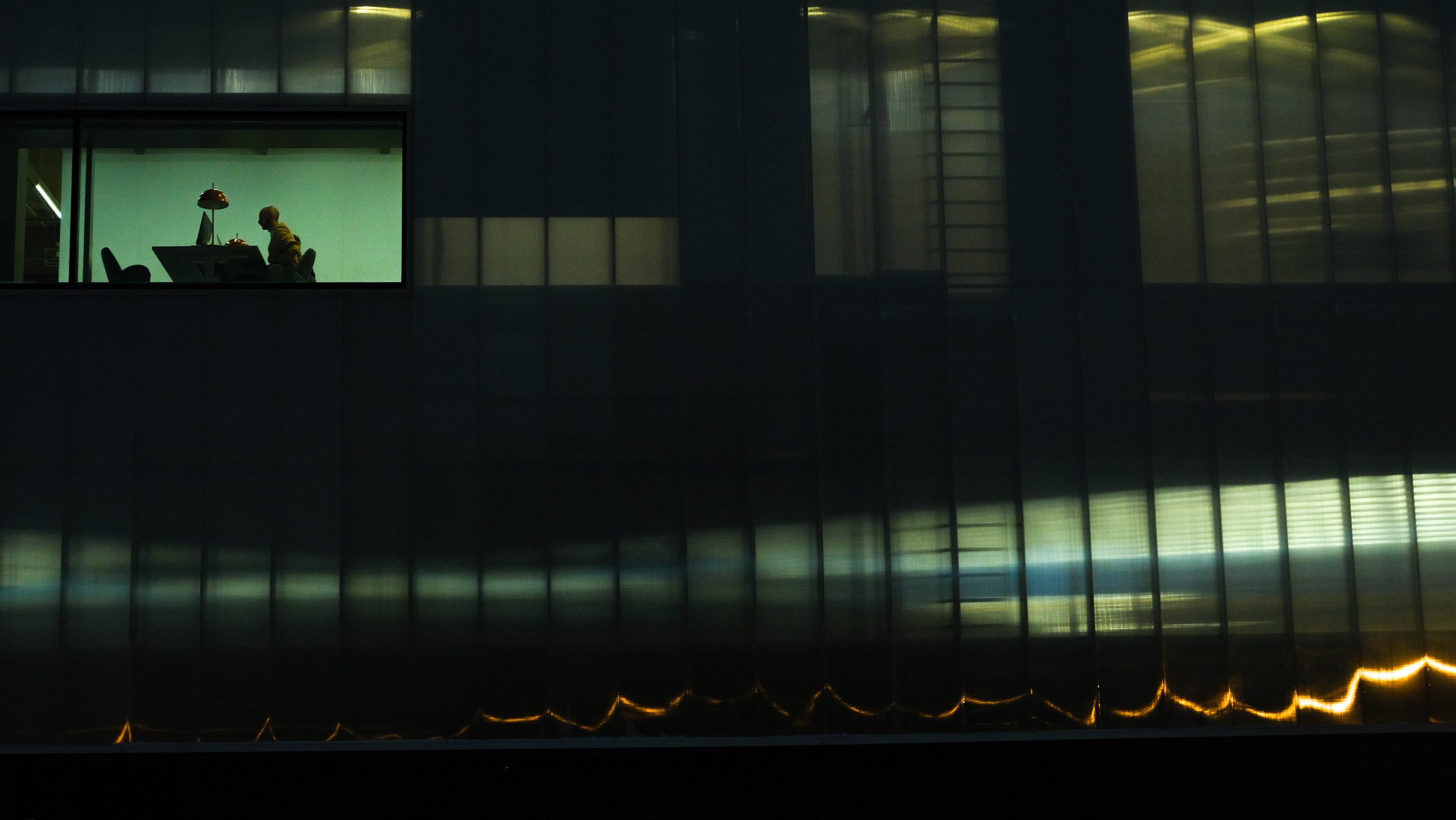 Silhouette of a person working at a desk in a brightly lit library room, viewed through a window from a dark exterior building at night. Cinematic photo scene with dramatic colors.