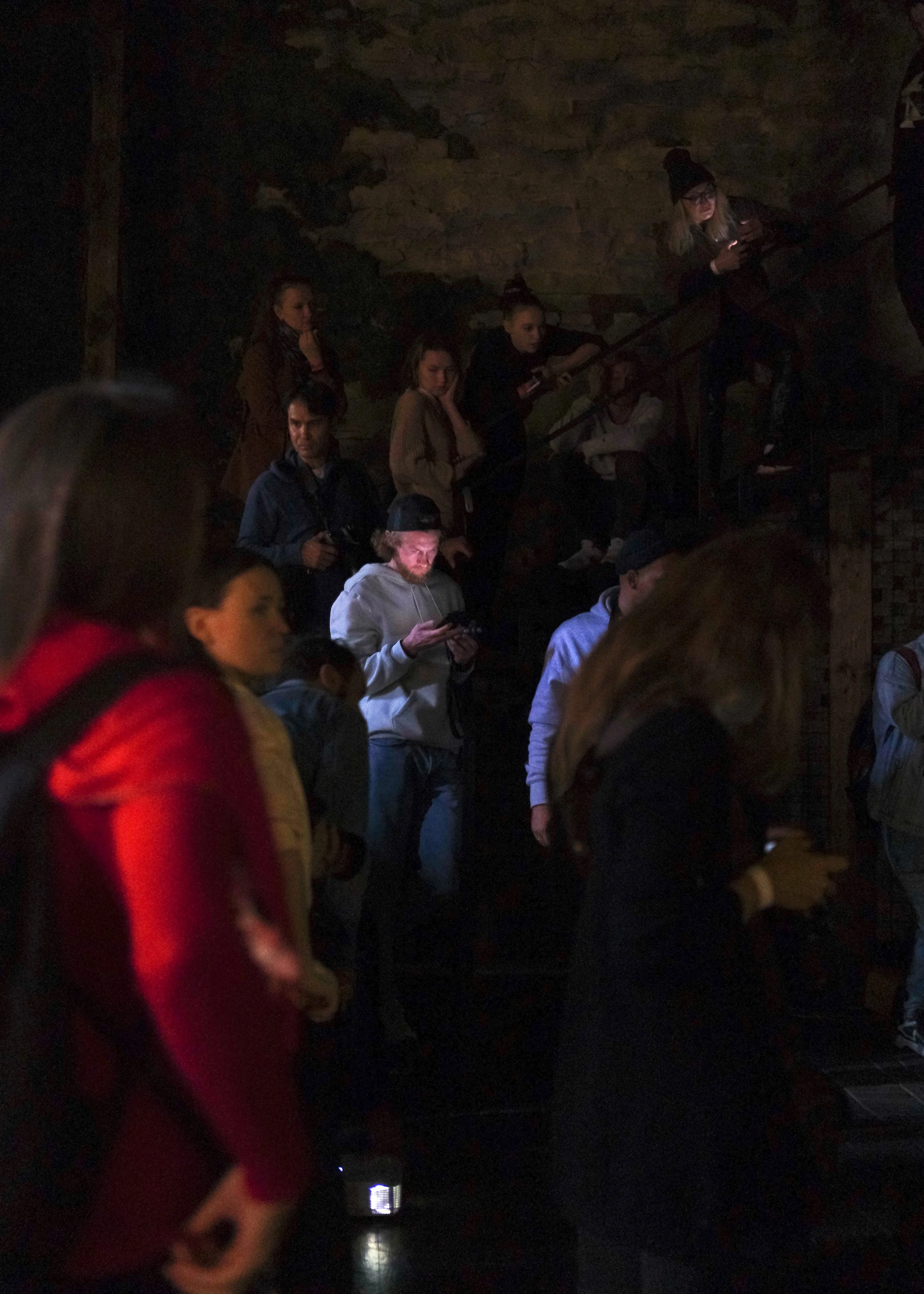 Group of people in a dark indoor setting, some looking at their phones, others observing surroundings.  A scene from ordinary life referring to the light and composition to Rembrandt's painting The Night Watch