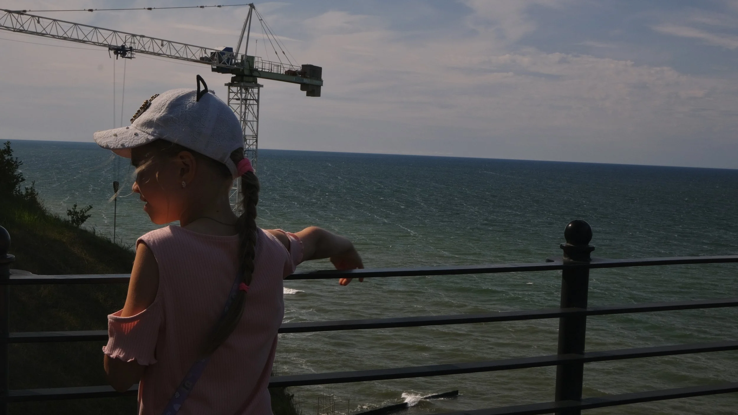 A young girl with a braid, wearing a pink shirt, a white cap, and a pink hair tie, leaning on a railing and pointing towards the ocean during daytime. A construction crane is visible in the background over the water.