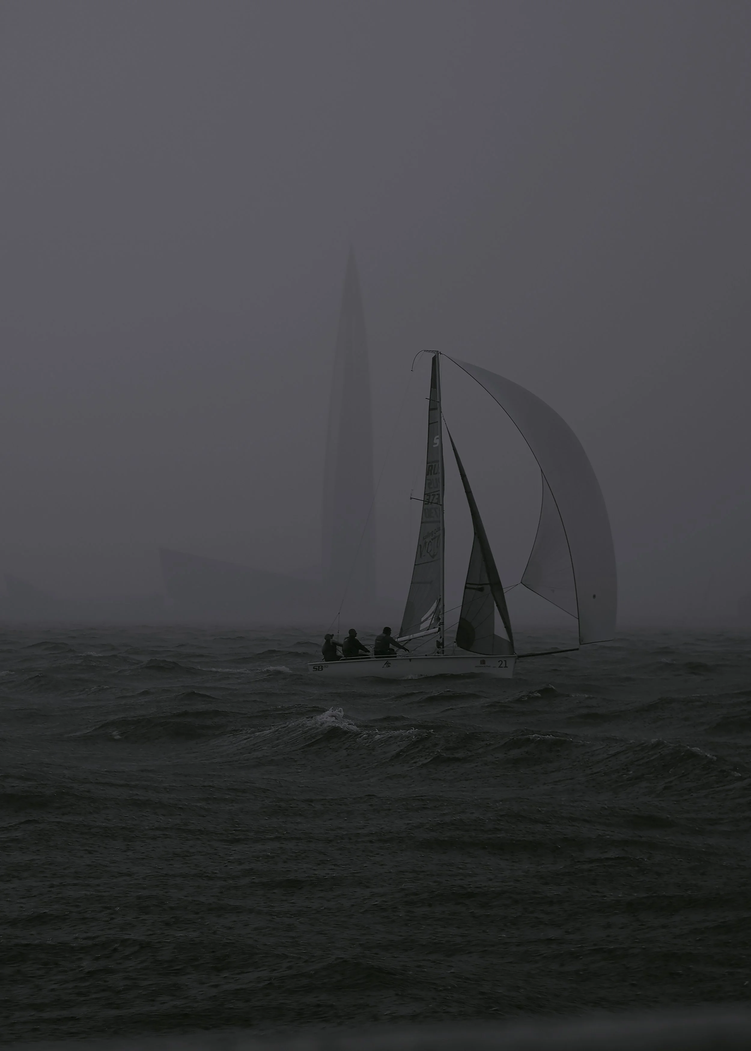 A sailboat with multiple sails navigating through rough waters on a foggy day.