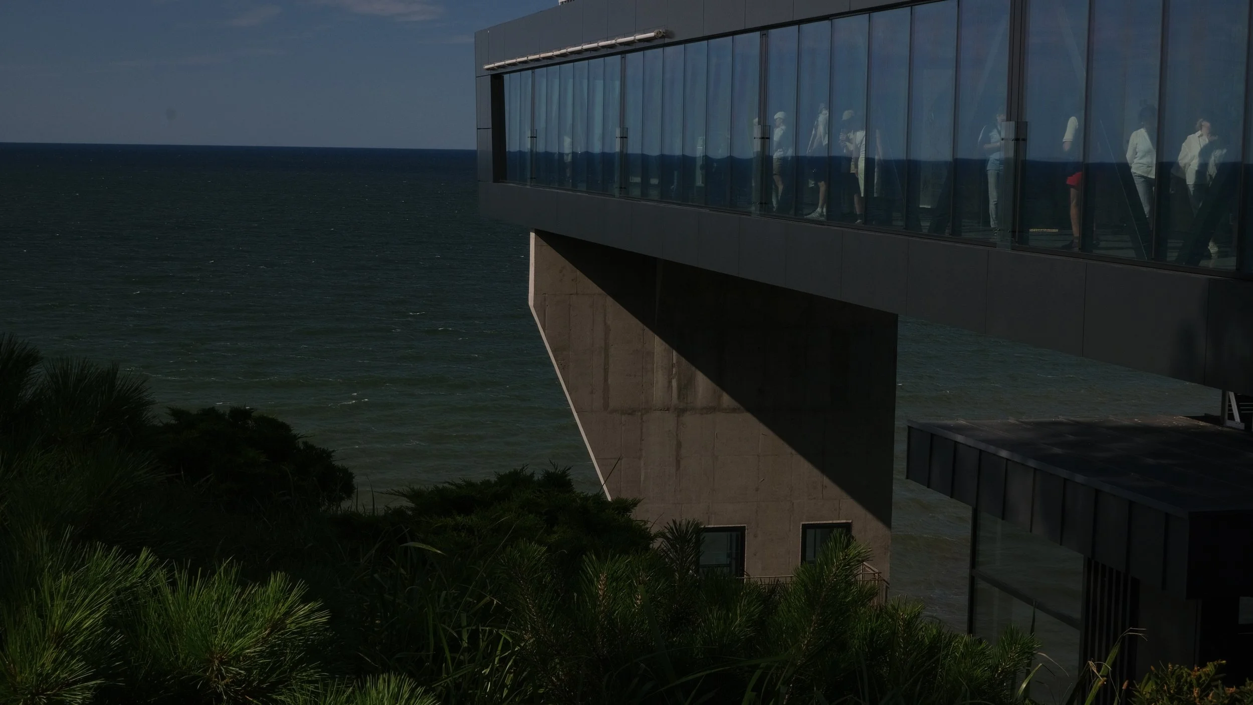 Modern building with a glass exterior overlooks the ocean, with some people visible inside.