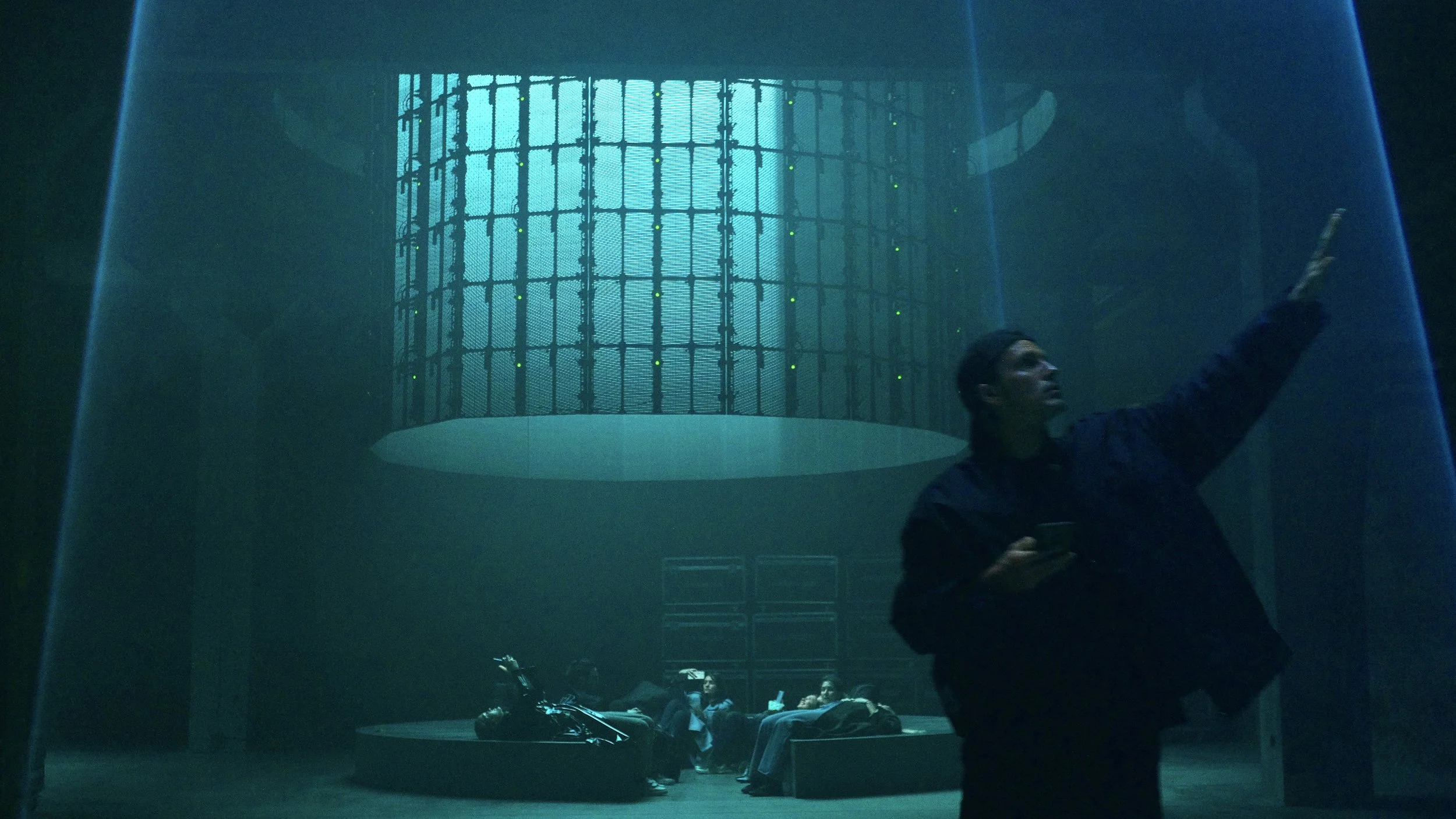 A man in a dark jacket holding a phone, pointing upward, with a large futuristic hardware or server structure and a group of people seated nearby in a dimly lit room.