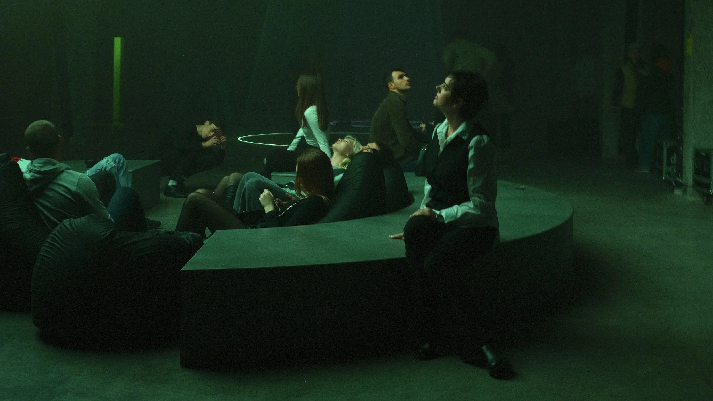 Group of people sitting and relaxing in a dark, green-lit room, some on bean bag chairs and some on a curved bench, with one person standing.