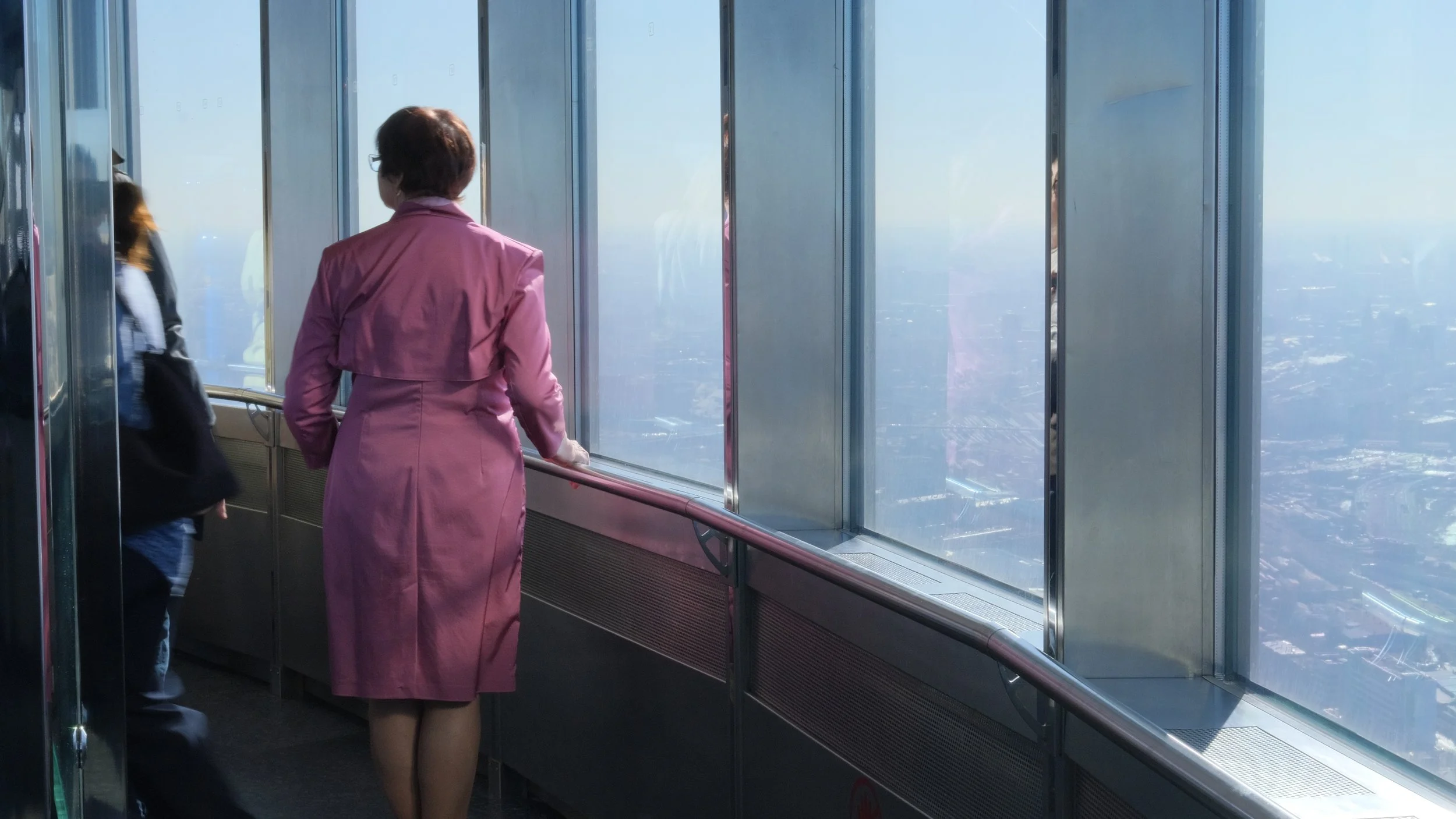 Woman in pink coat looking out the window from a high observation deck with a cityscape below.