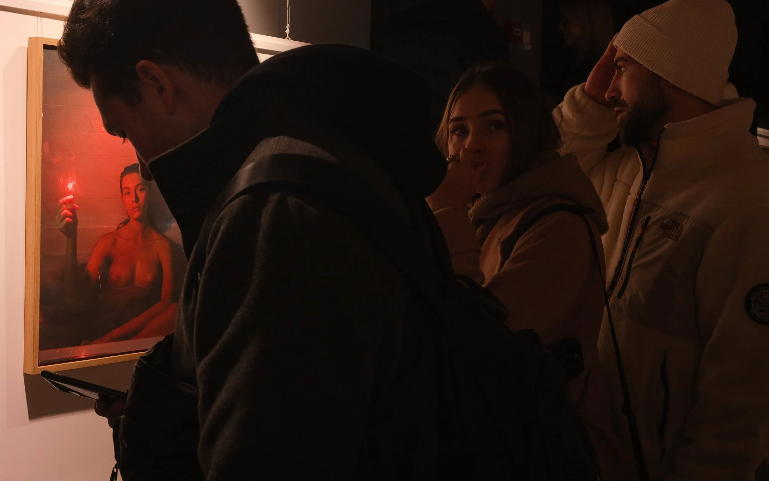 People viewing a framed photo of a topless woman holding a red light in an art gallery.