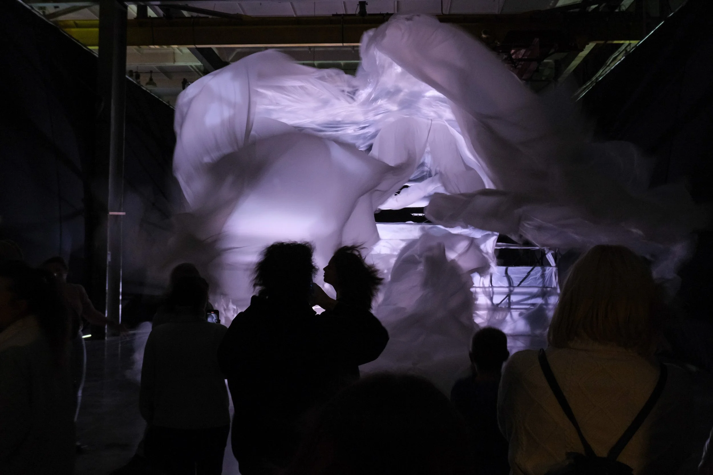 People observing an illuminated art installation featuring flowing white fabric in a dark indoor space.