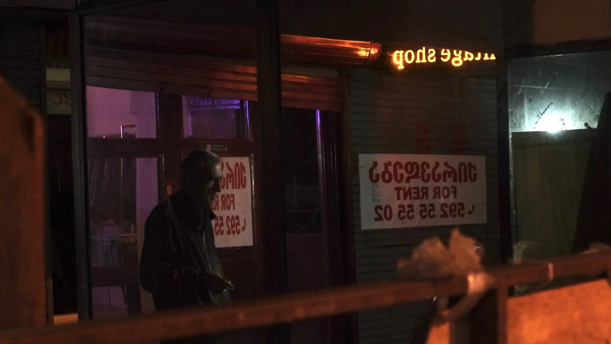 A person standing inside a dimly lit storefront or building at night, looking outside through glass, with signs visible on the window and an orange illuminated sign above.