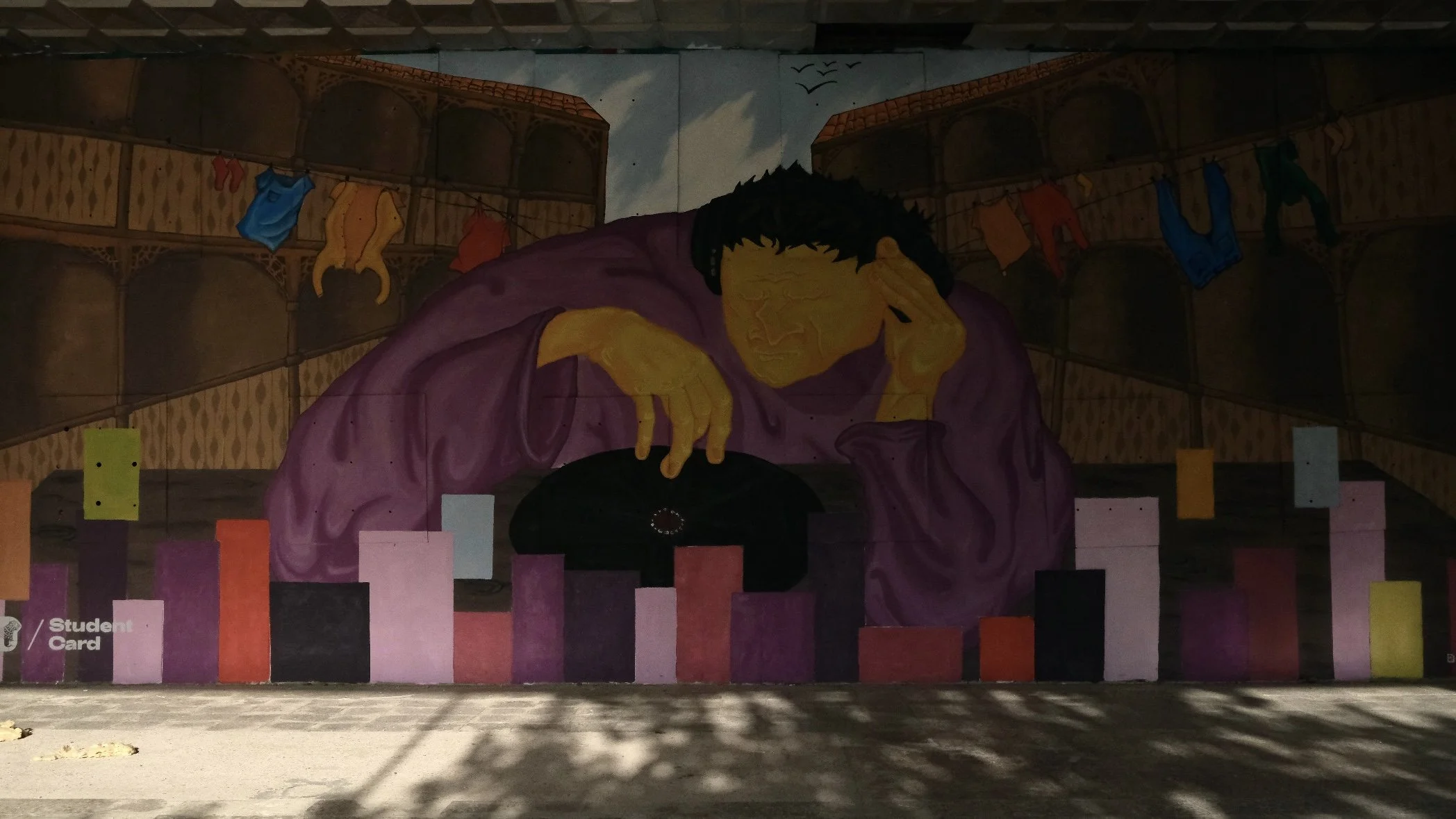 Street art mural of a person with yellow skin and black hair, wearing a purple shirt, resting their head on their hand, with colorful flags hanging in the background.