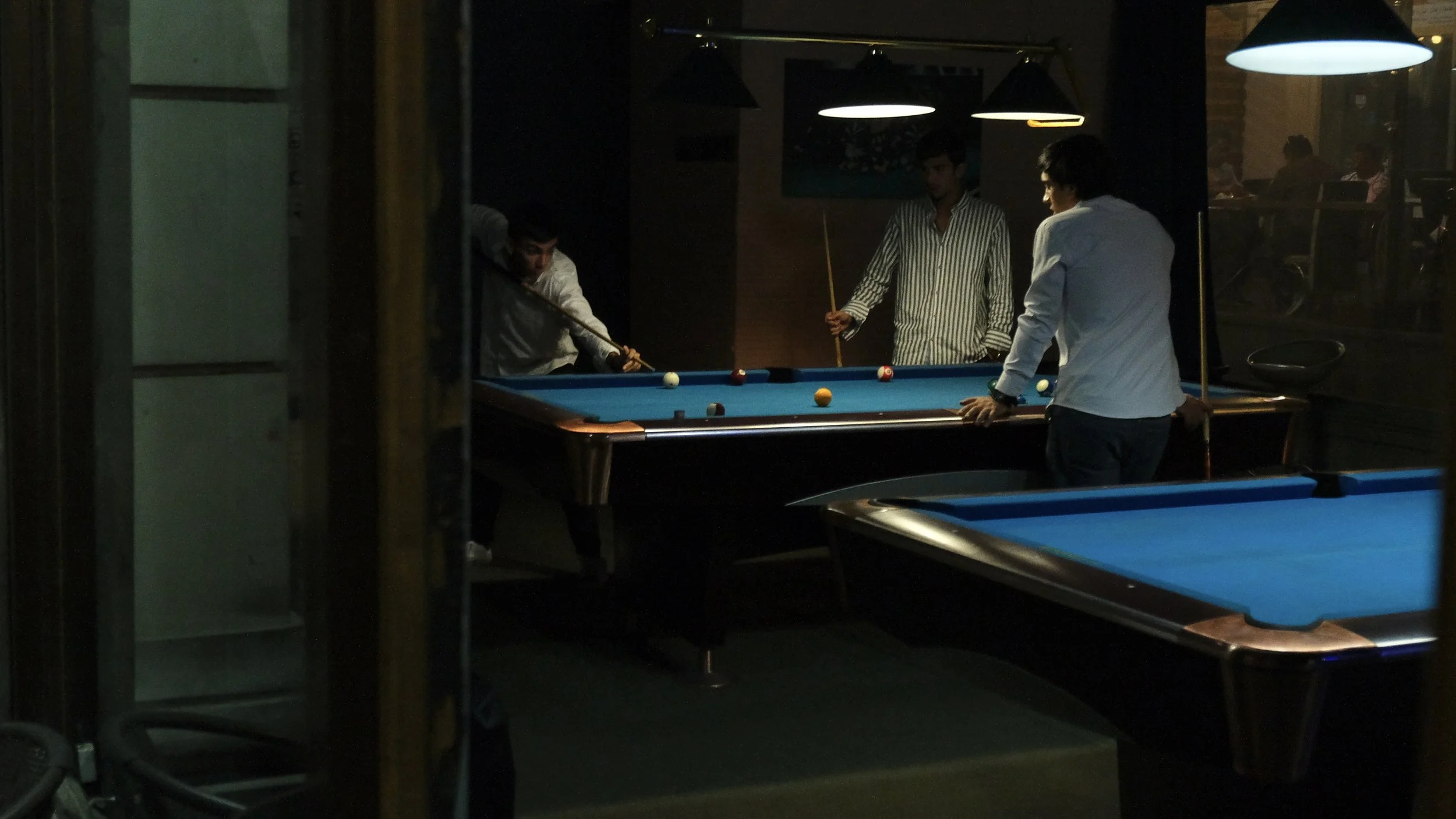 Four men playing billiards in a dimly lit pool hall, with overhead lamps illuminating the tables.