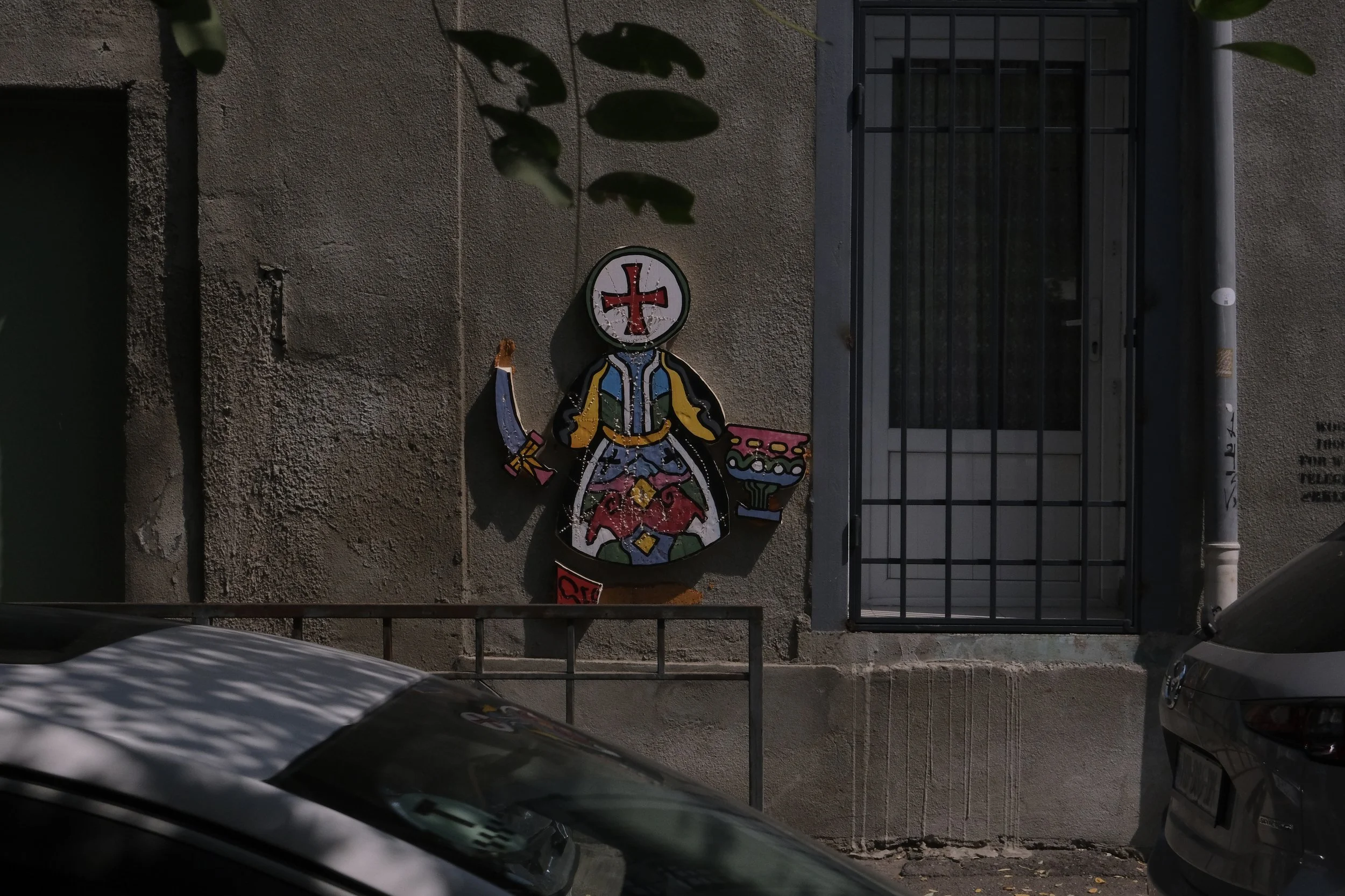 Colorful graffiti mural of a stylized woman with a mask and a hammer, standing beside a pink bowl, painted on a gray building wall next to a barred window.