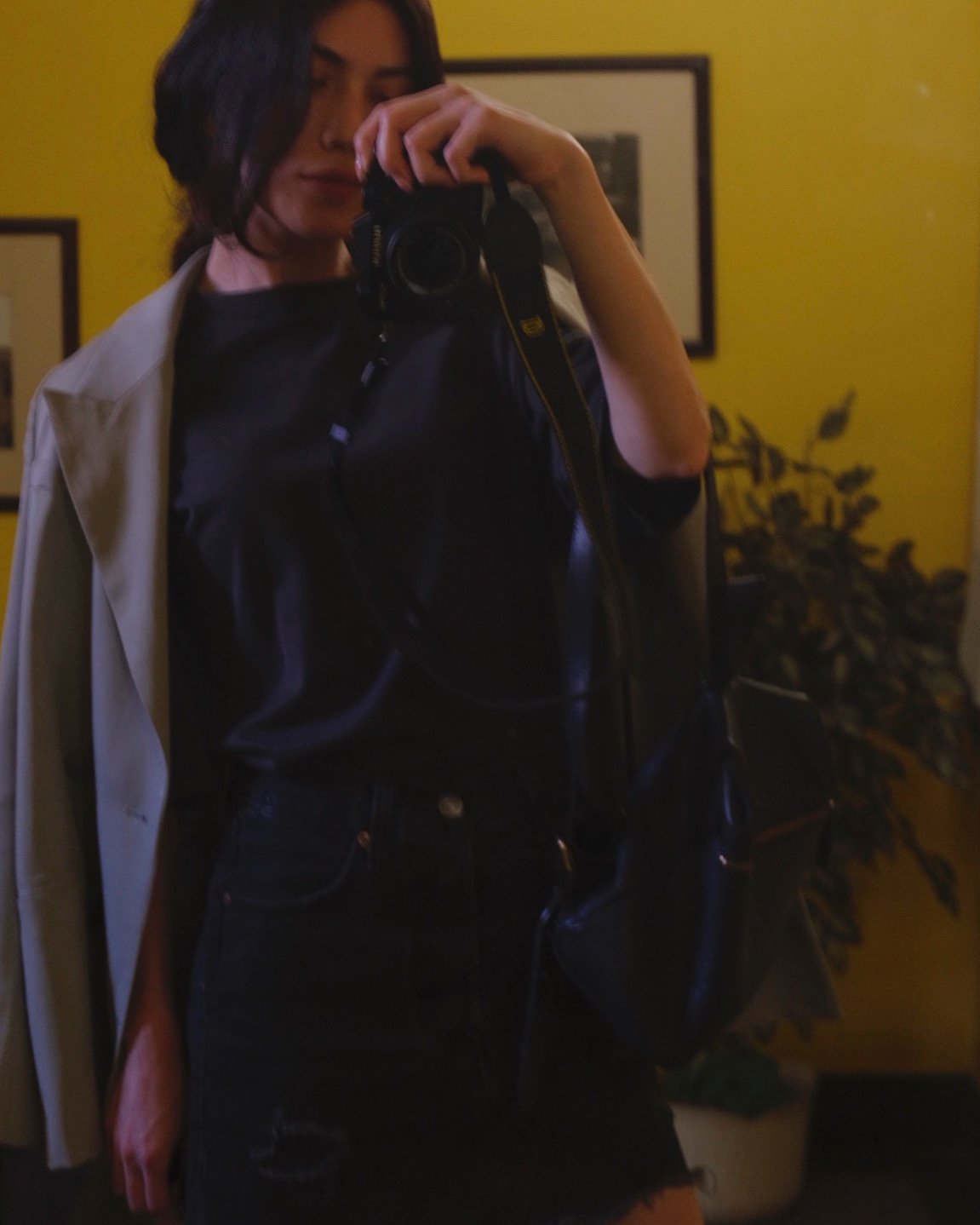A woman taking a mirror selfie with a camera, wearing a black top, black ripped jeans, and a light-colored coat draped over her shoulders. She has dark hair and a neutral expression, with framed artwork and a plant in the background.