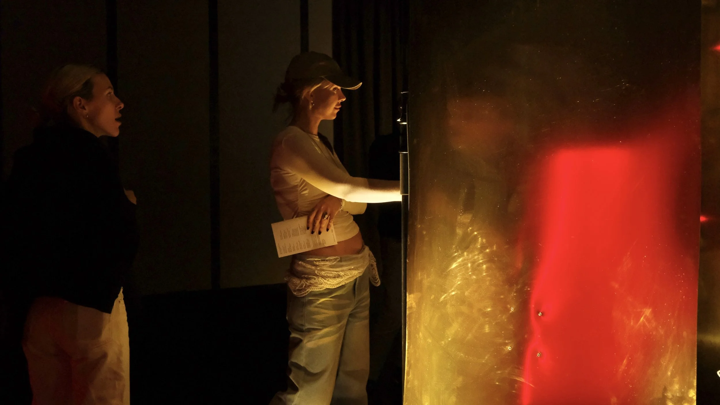 Two women standing in front of an illuminated, reflective art installation with red and gold colors.
