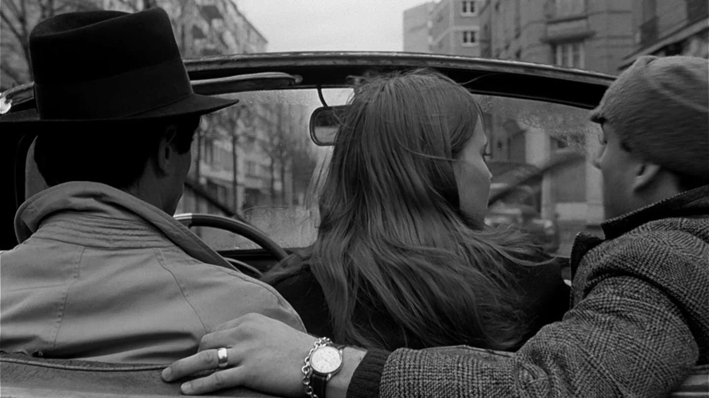 Three people sitting in a car, with buildings visible through the windshield; one person wearing a hat, another with long hair, and a third in a cap and patterned jacket, appearing to be engaged in conversation.