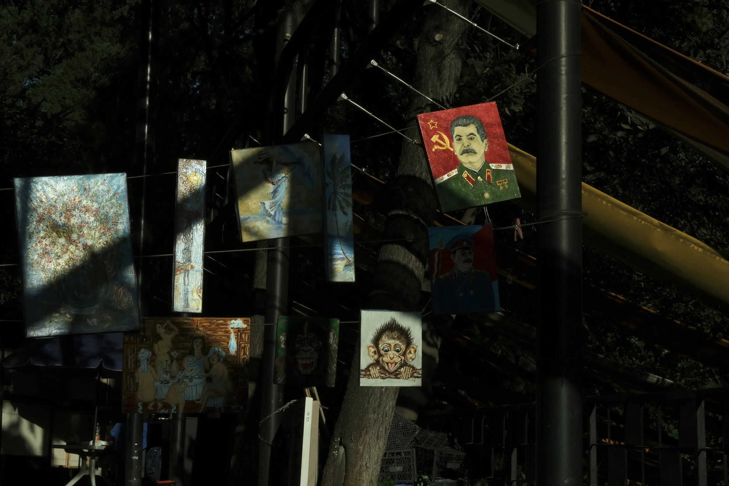 Various colorful paintings and posters hanging on strings tied around a tree and metal poles, including portraits of Soviet leaders and artistic scenes.