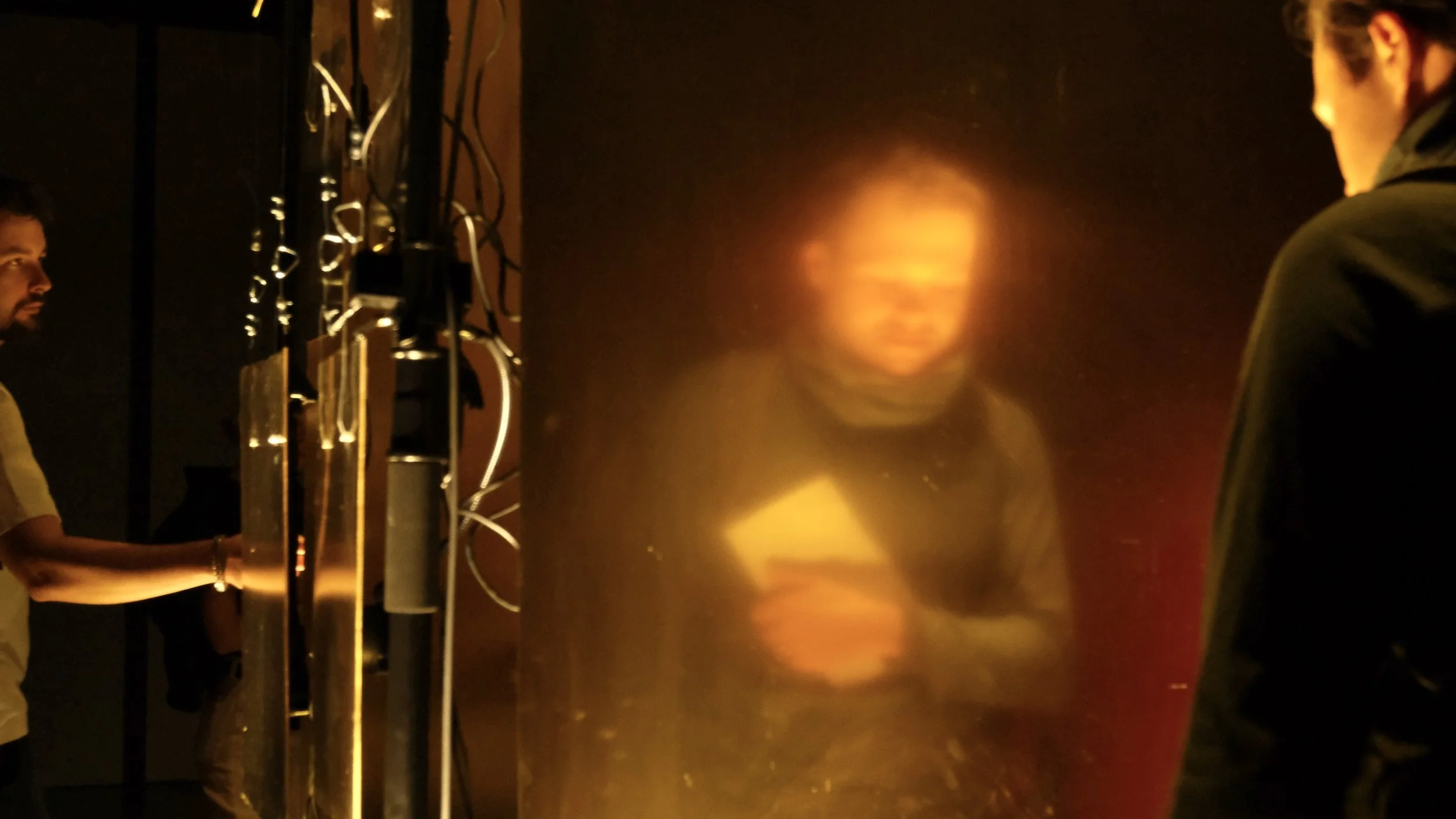A blurry woman holding a phone, surrounded by two men in dark clothing, in a dimly lit environment with orange hues and electronic equipment.