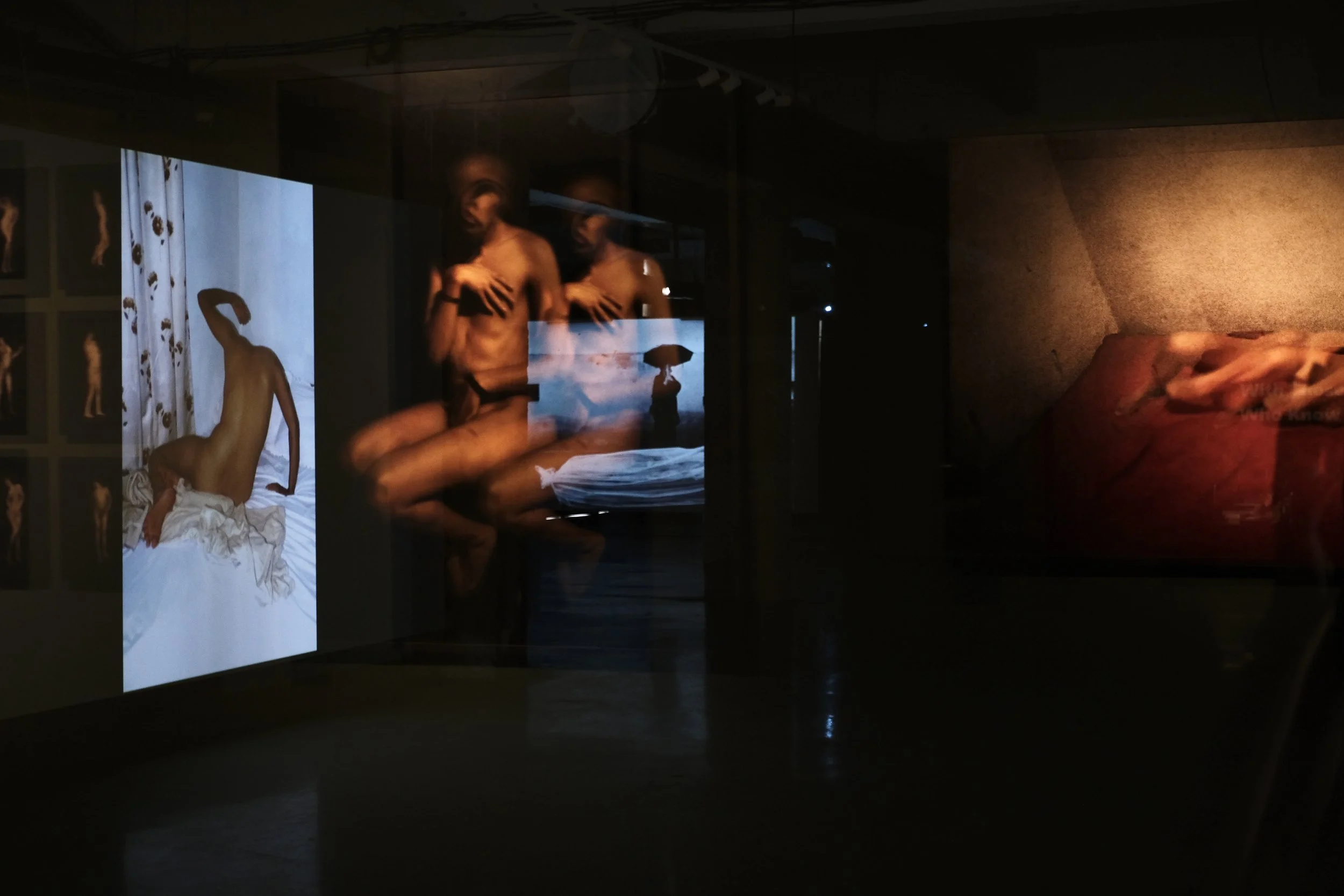 Dark room with digital art projections showing a nude figure on a bed and close-up of a person holding an umbrella near the ocean.