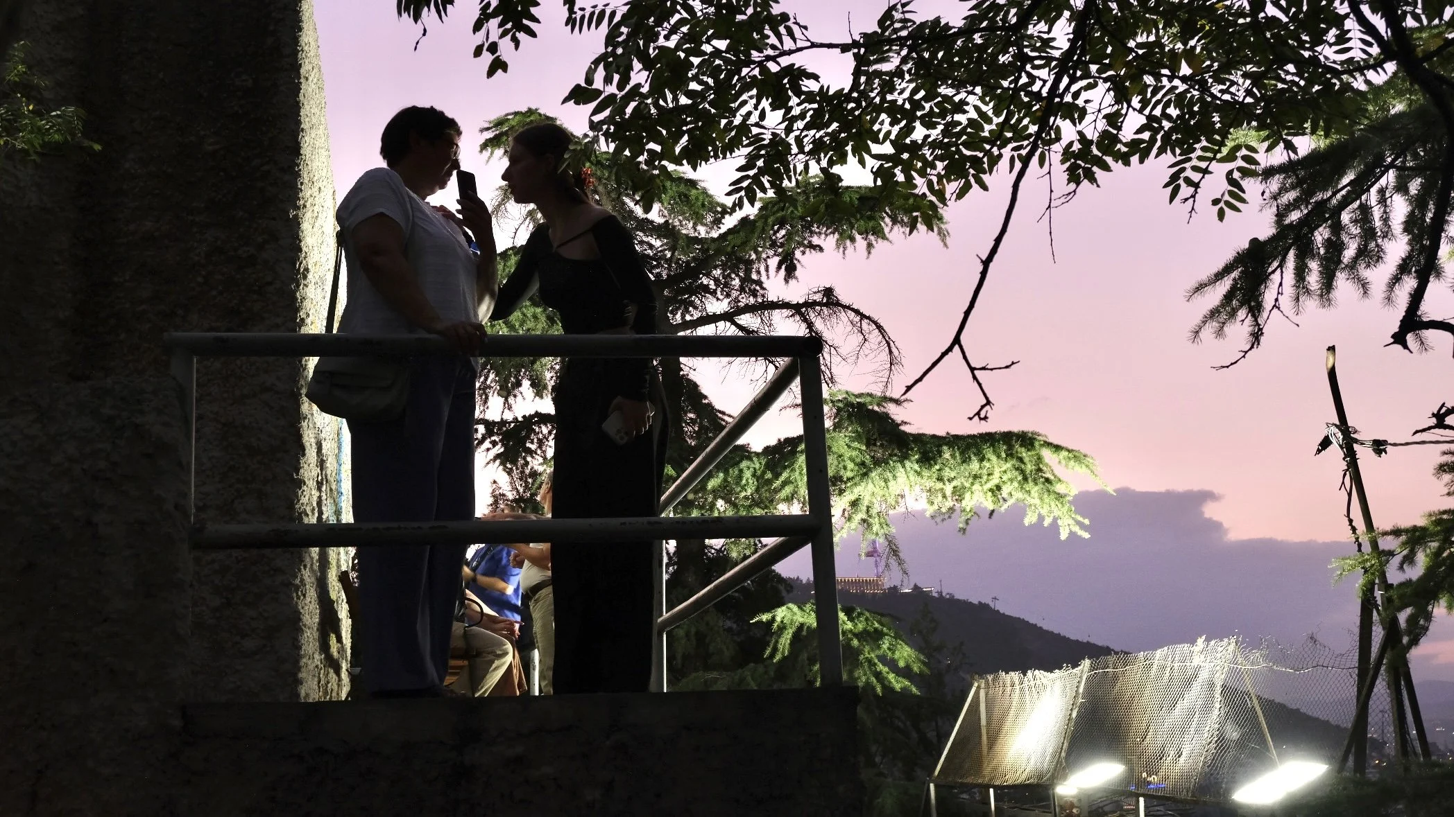 Silhouettes of two women talking on a balcony during sunset with trees and hills in the background.