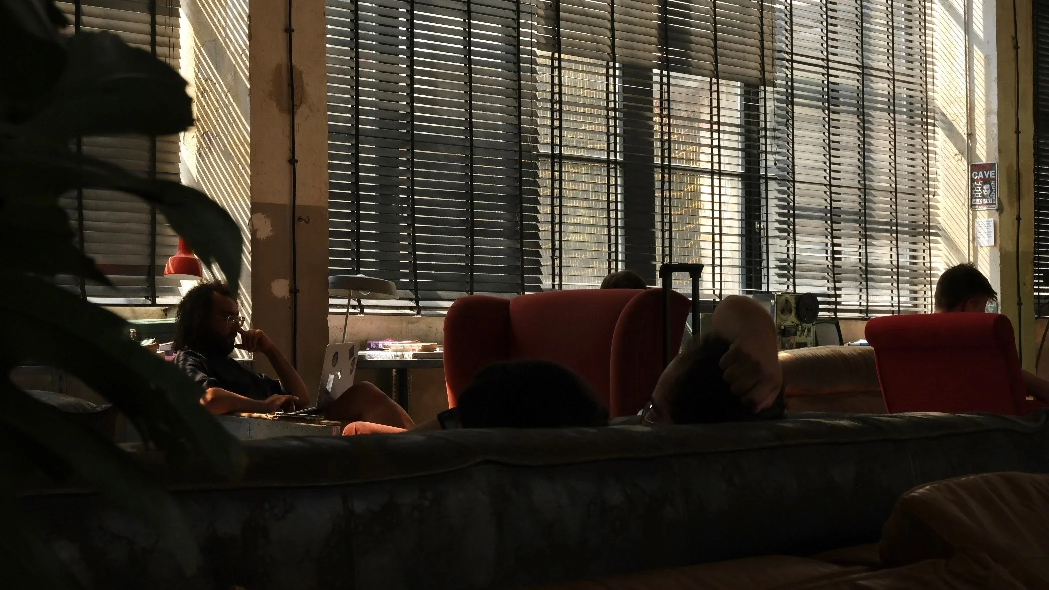 People sitting in a dimly lit lounge area with large windows and horizontal blinds, some using laptops, with a relaxed ambiance.