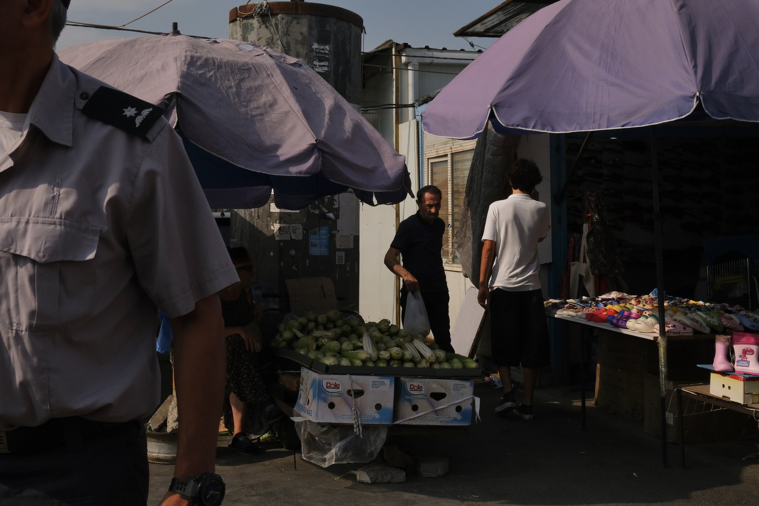 Street market scene with vendors selling vegetables and shoes under umbrellas, with a person wearing a uniform on the left side. The action takes place at a market in Tbilisi.