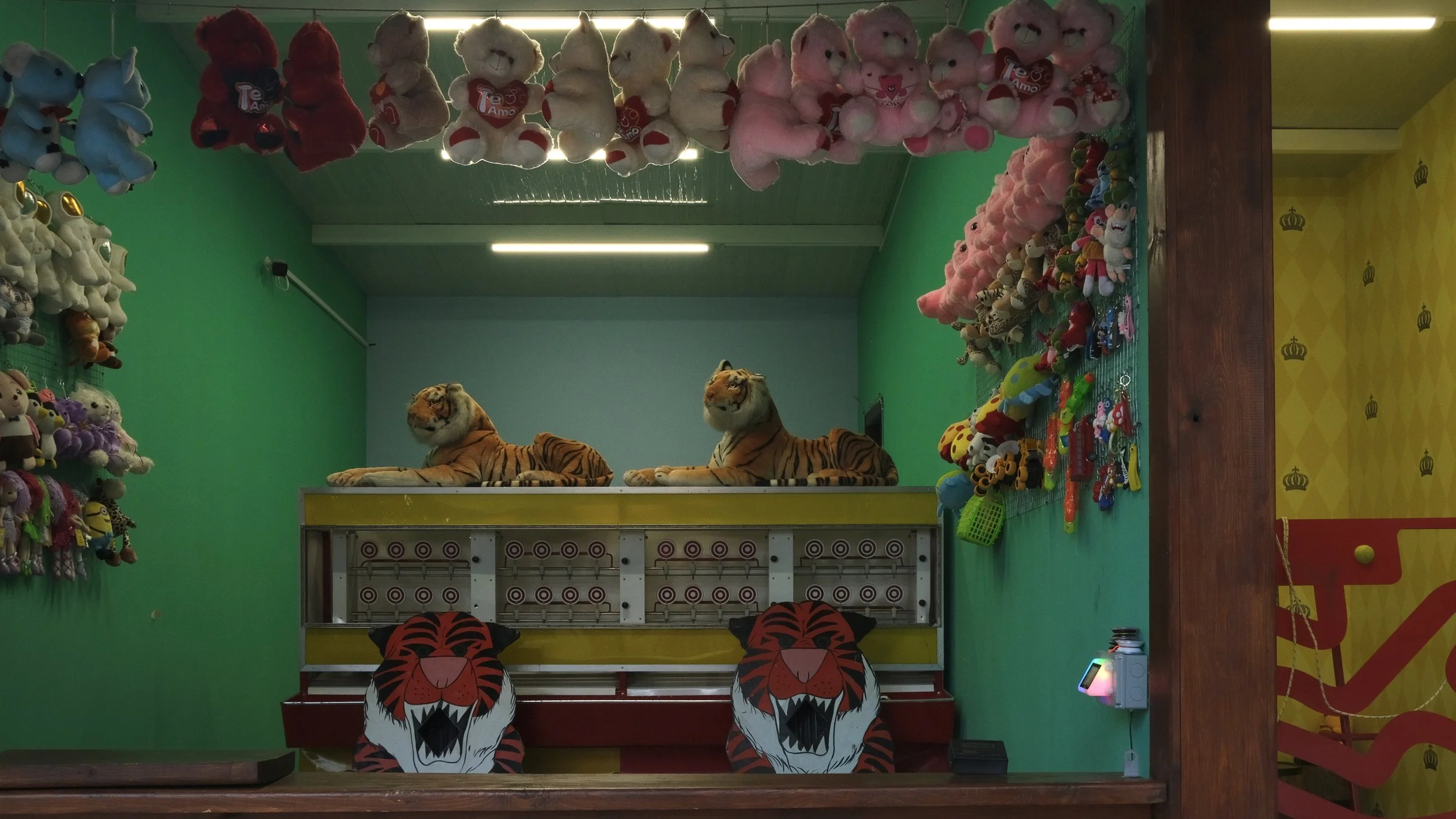 A colorful amusement arcade with plush toys, including stuffed animals hanging from the ceiling, and tiger plush toys on a platform. There are decorative tiger faces on the barrier in front, and the walls are painted green with a yellow section on th
