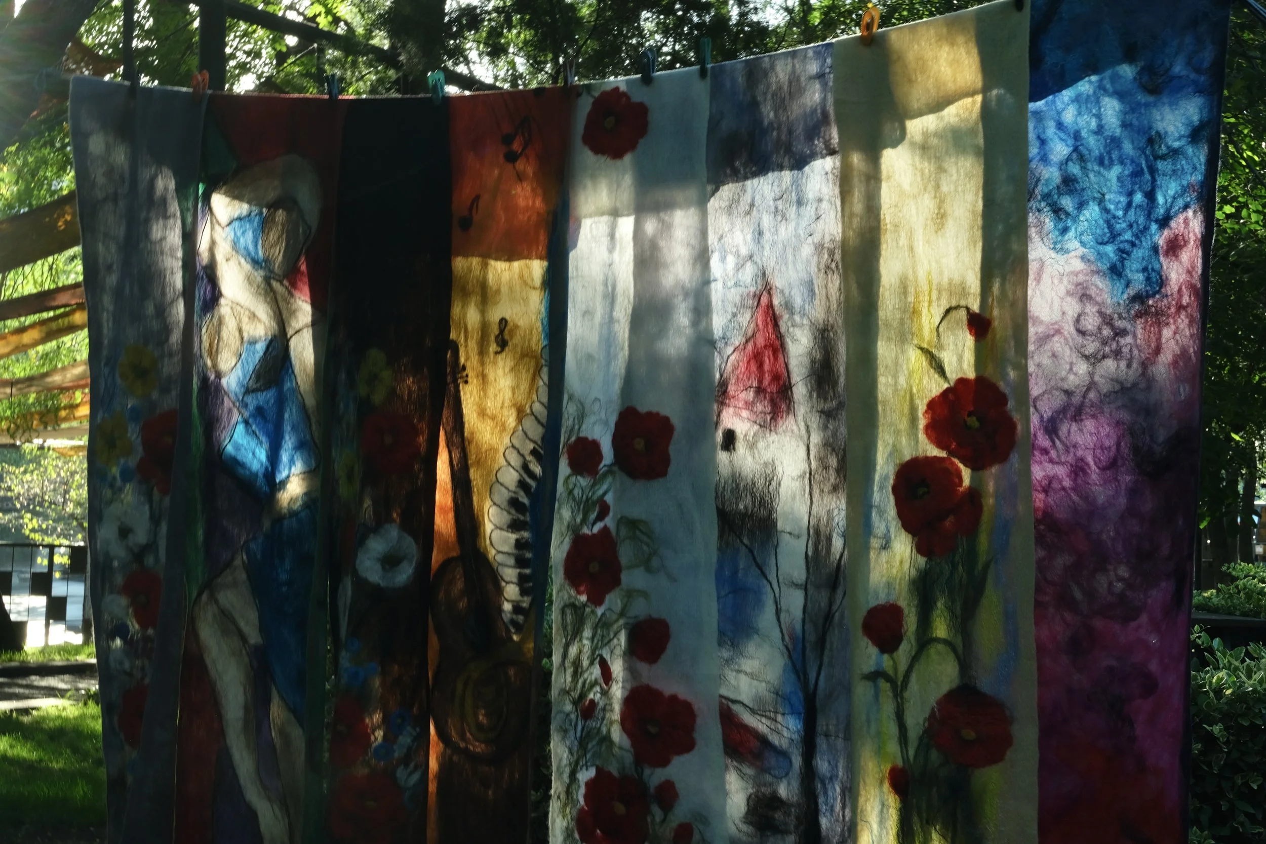 Colorful fabric or paper panels hanging outdoors, decorated with abstract art, red flowers, and musical notes, with trees and sunlight in the background.