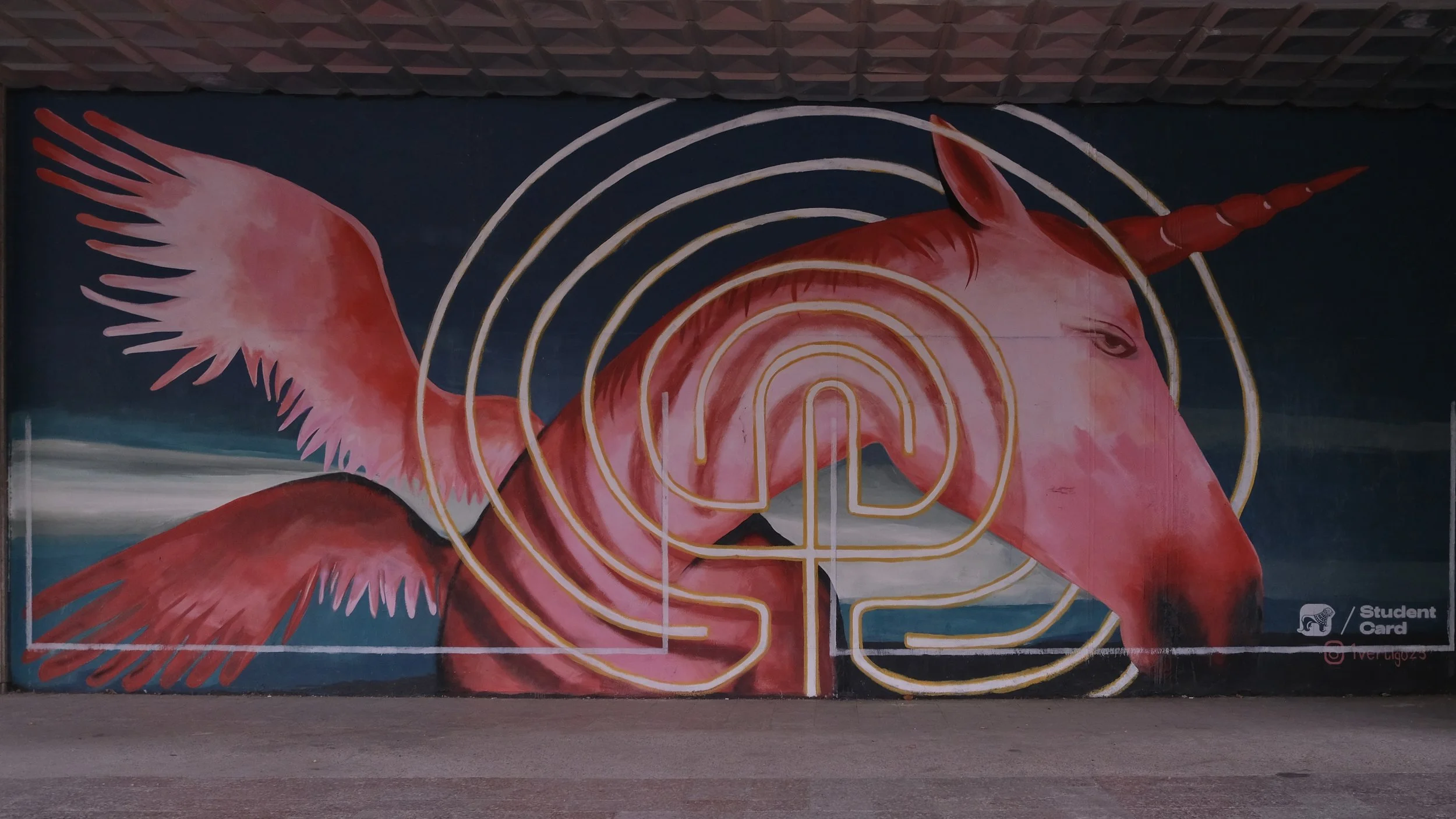 A mural of a pink unicorn with wings, a spiraling tail, and a golden maze pattern on its face, with concentric circles around its head, on a dark background.