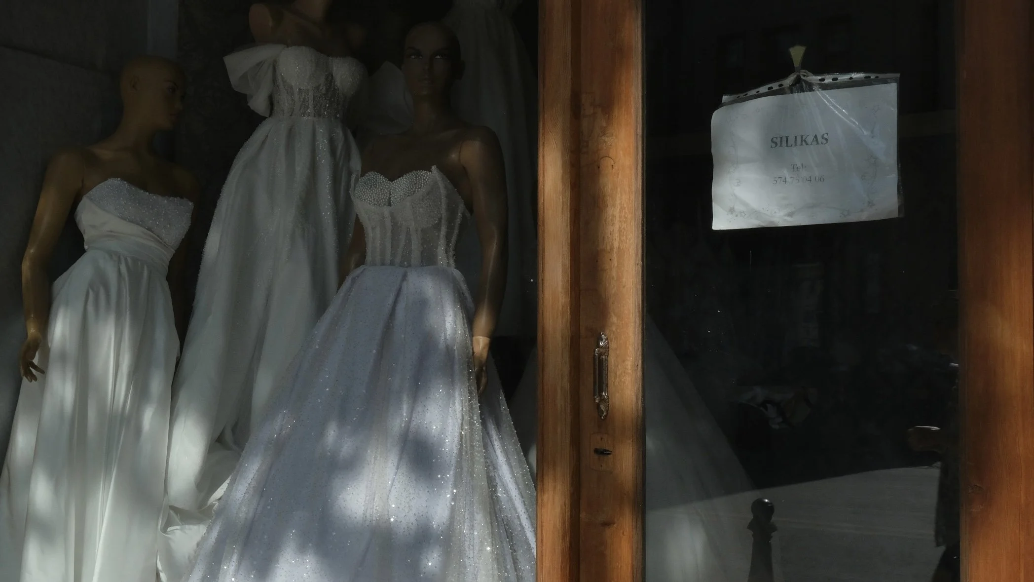 Storefront window displaying mannequins dressed in white wedding gowns, with a sign on the side labeled 'SILIKAS' and a phone number.