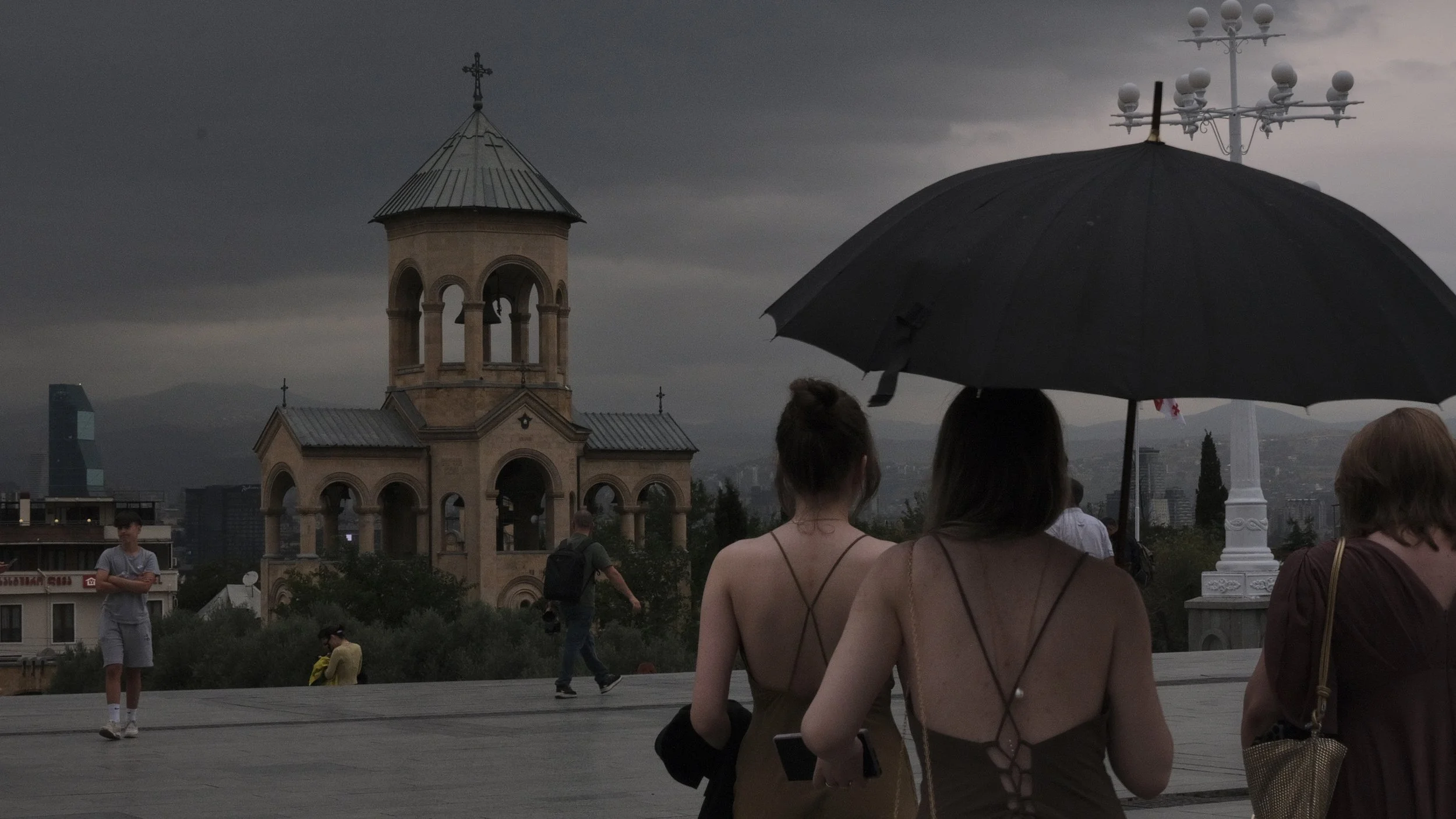 Women sitting on a rooftop under an umbrella, with a church and cityscape in the background on a cloudy day.