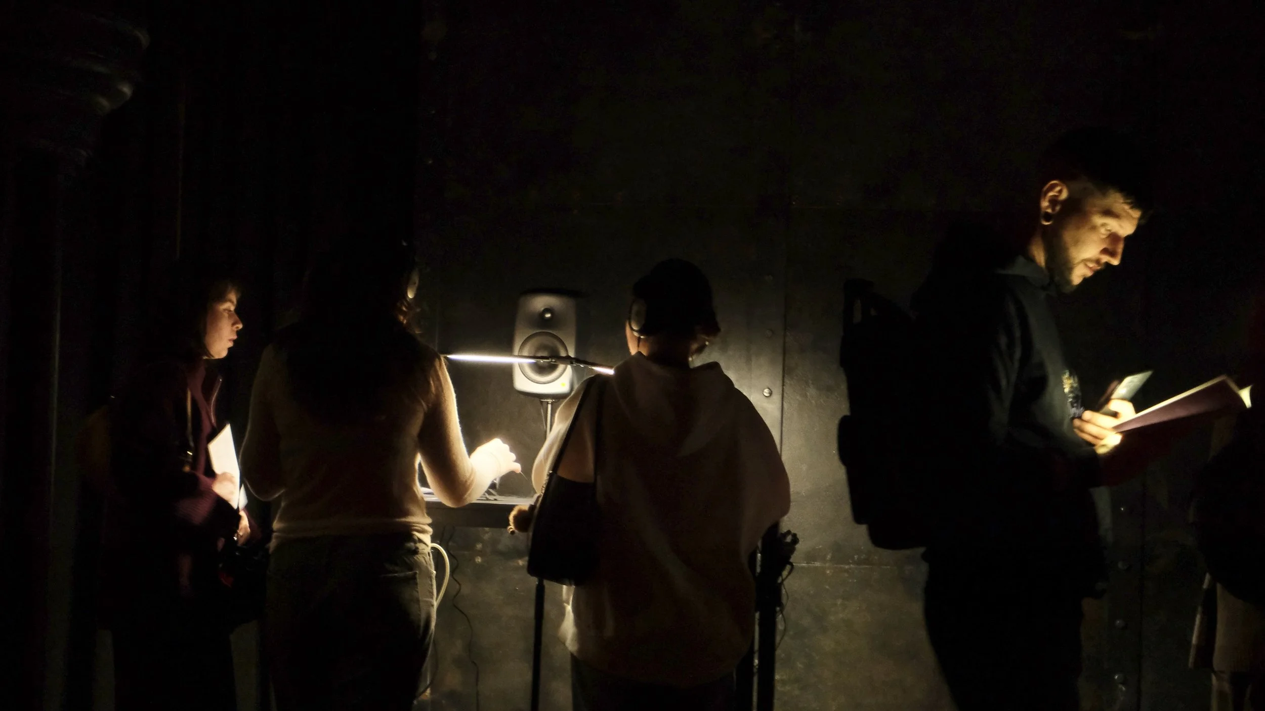 People standing in a dark room, illuminated by a small amount of light, some reading or holding papers.