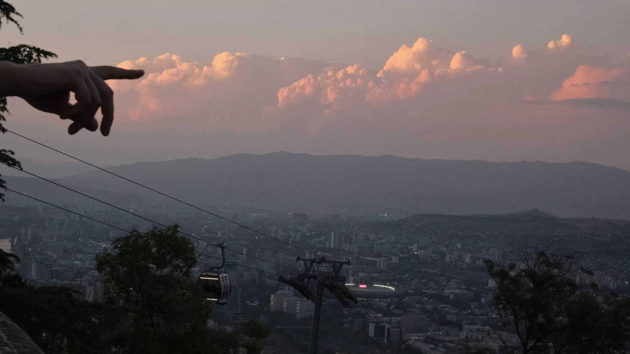 A person's hand pointing towards pink and orange clouds in the sky over a cityscape with mountains in the background, and a cable car and power lines in the foreground.