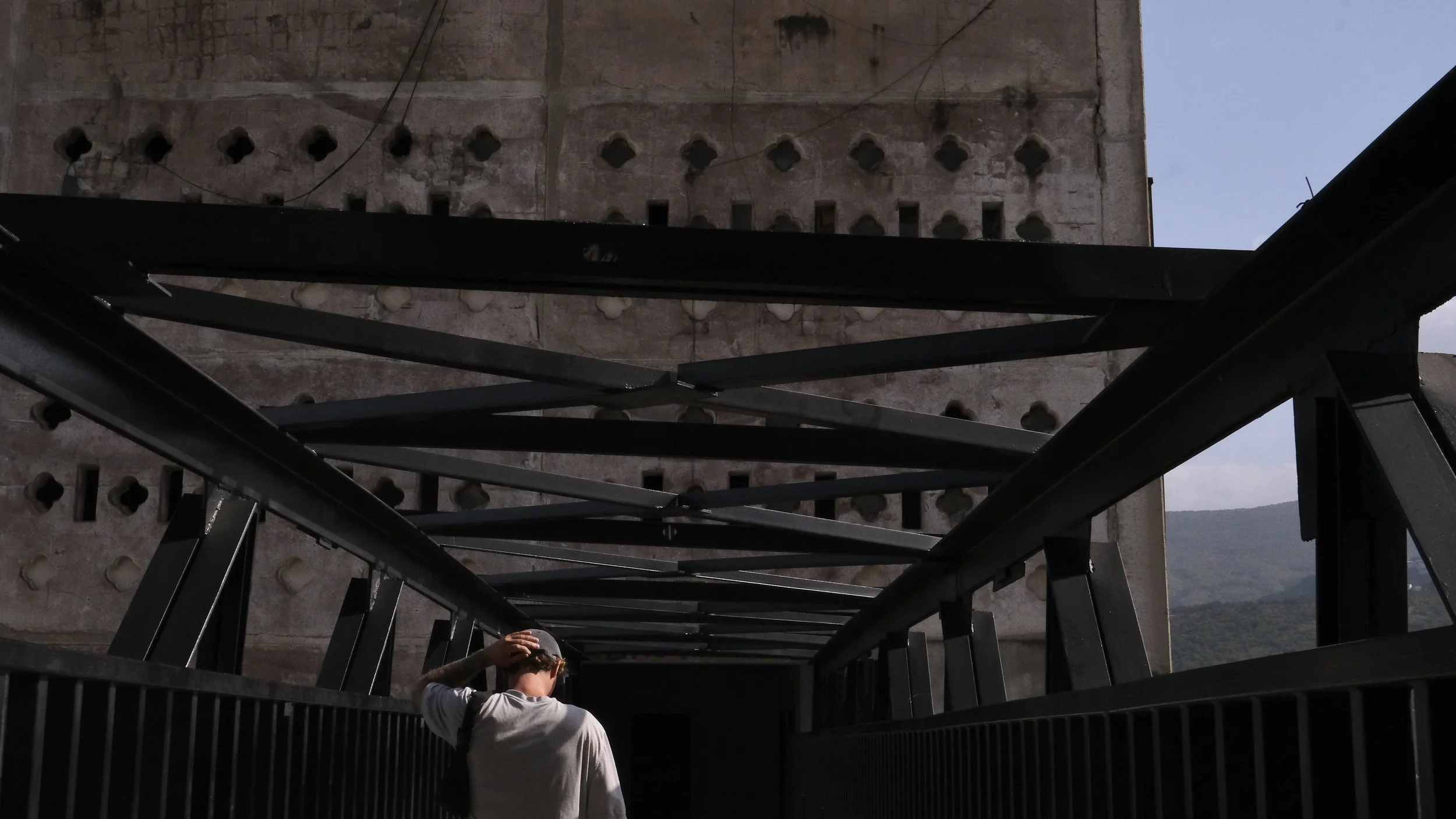 Person walking on a bridge walkway under a large, old concrete structure with perforated patterns, with mountains in the background