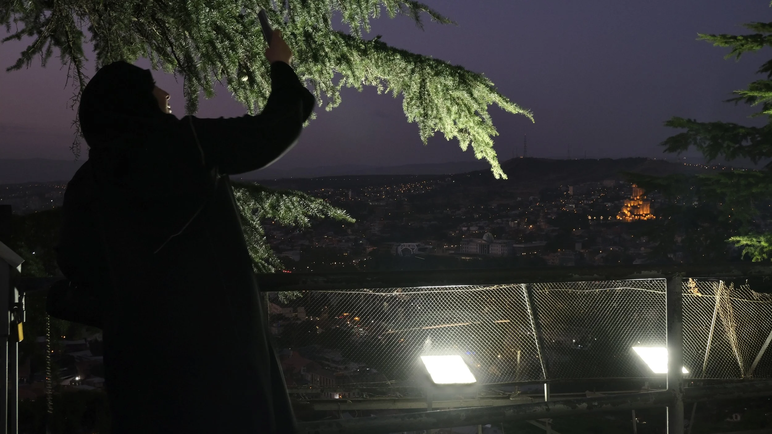 A woman taking a photo of a city skyline at night from a lookout point with a metal railing and bright lights, surrounded by trees.