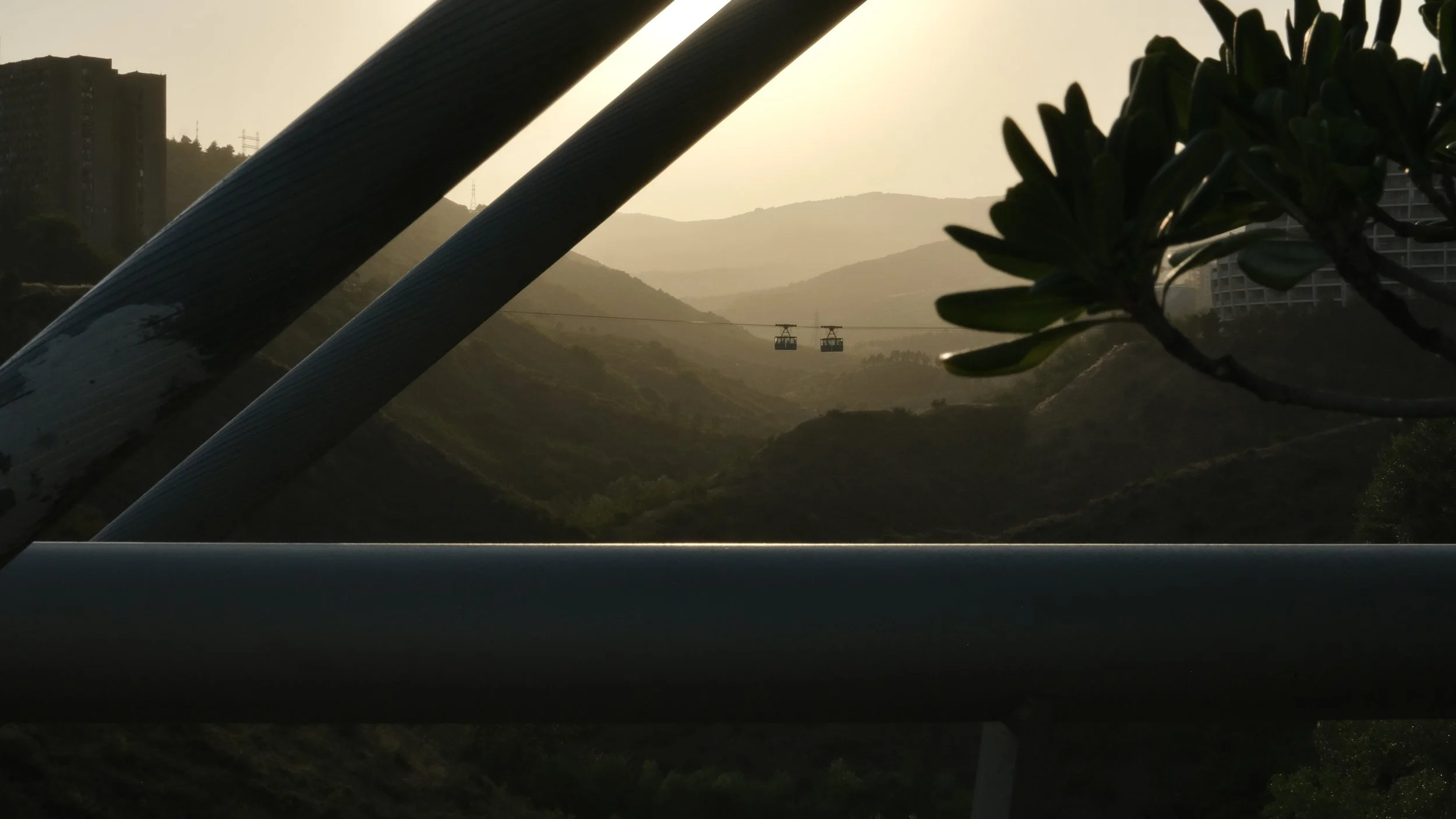 A scenic view of mountains at sunset with two cable cars hanging in the distance, framed by a tree branch and large beams in the foreground.