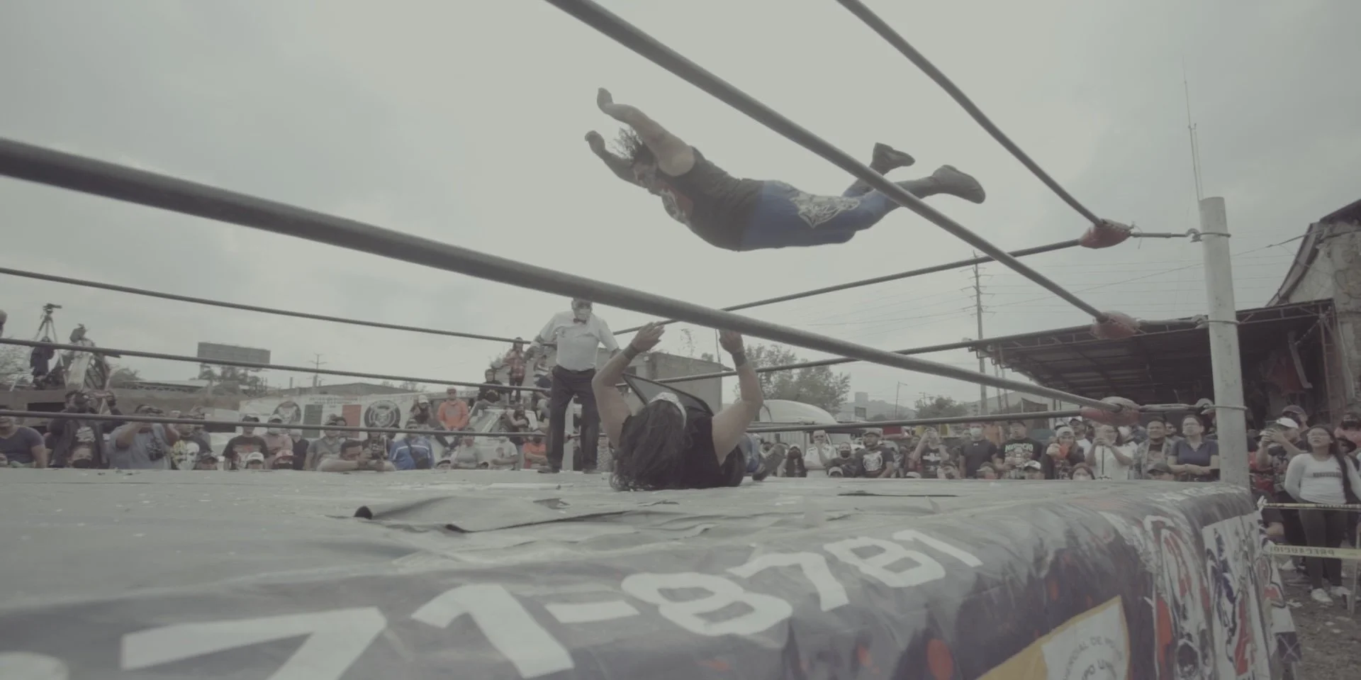 A wrestling match taking place outdoors with a crowd watching. One wrestler is on the mat, and another is flying through the air towards him.