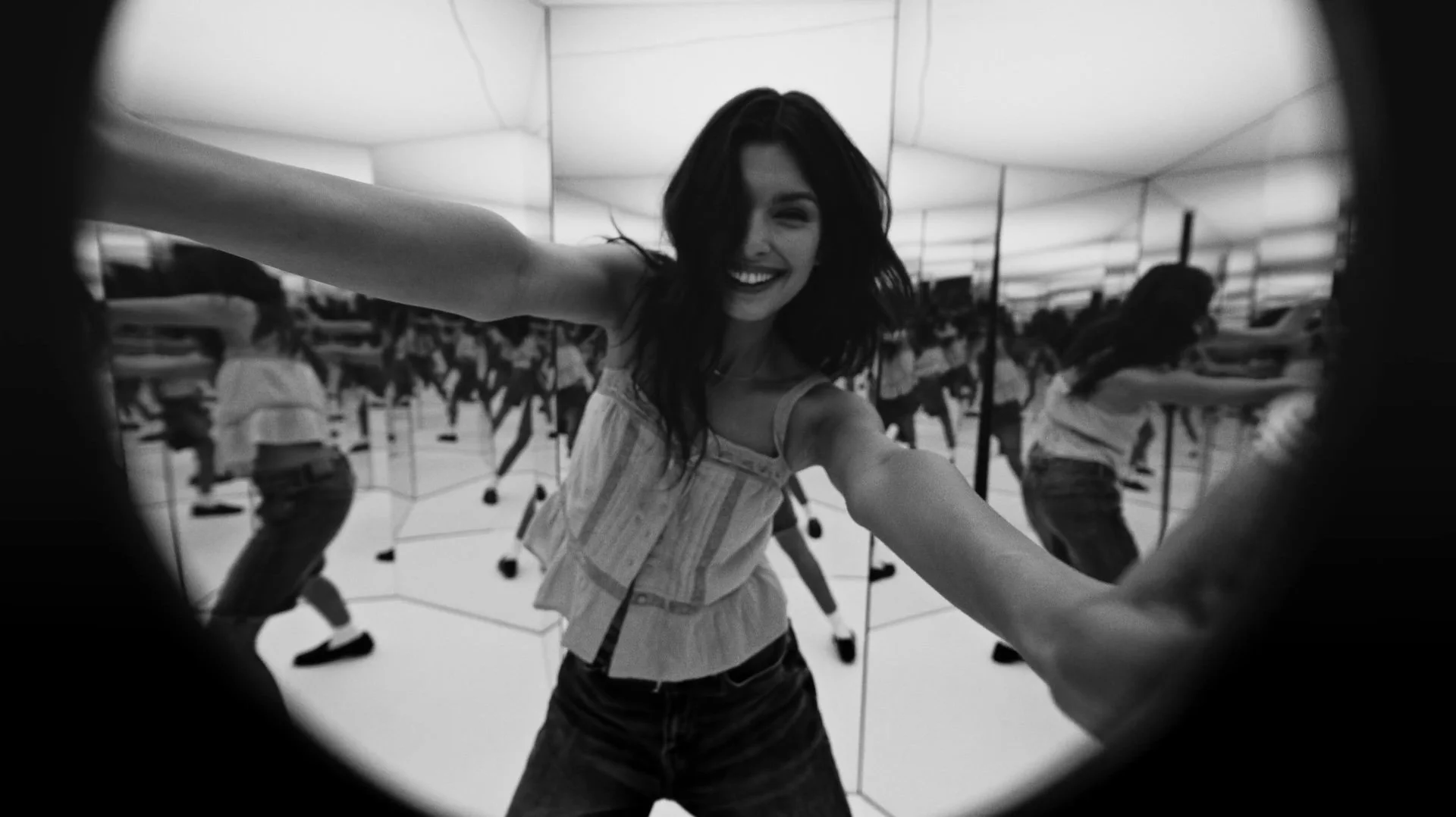 Black and white photo of a woman taking a selfie in a room with mirrors, smiling and stretching out her arms, with multiple people reflected in the mirrors in the background.