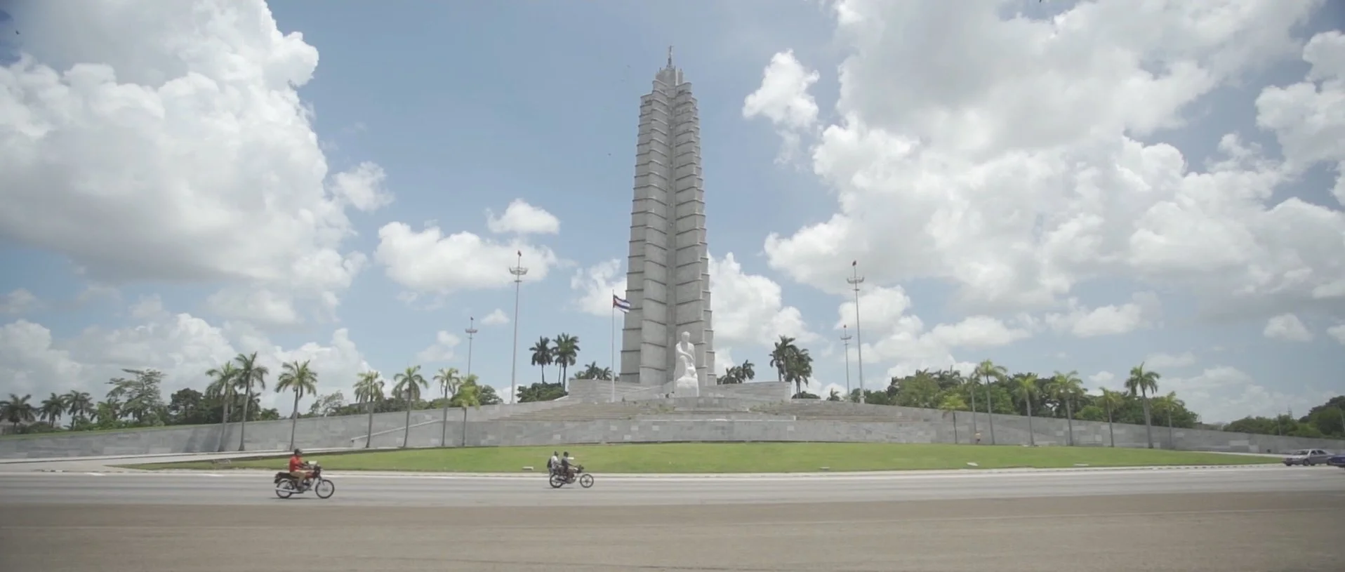A tall white monument with a statue at the base, surrounded by palm trees and a road with motorcyclists passing by, under a partly cloudy sky.