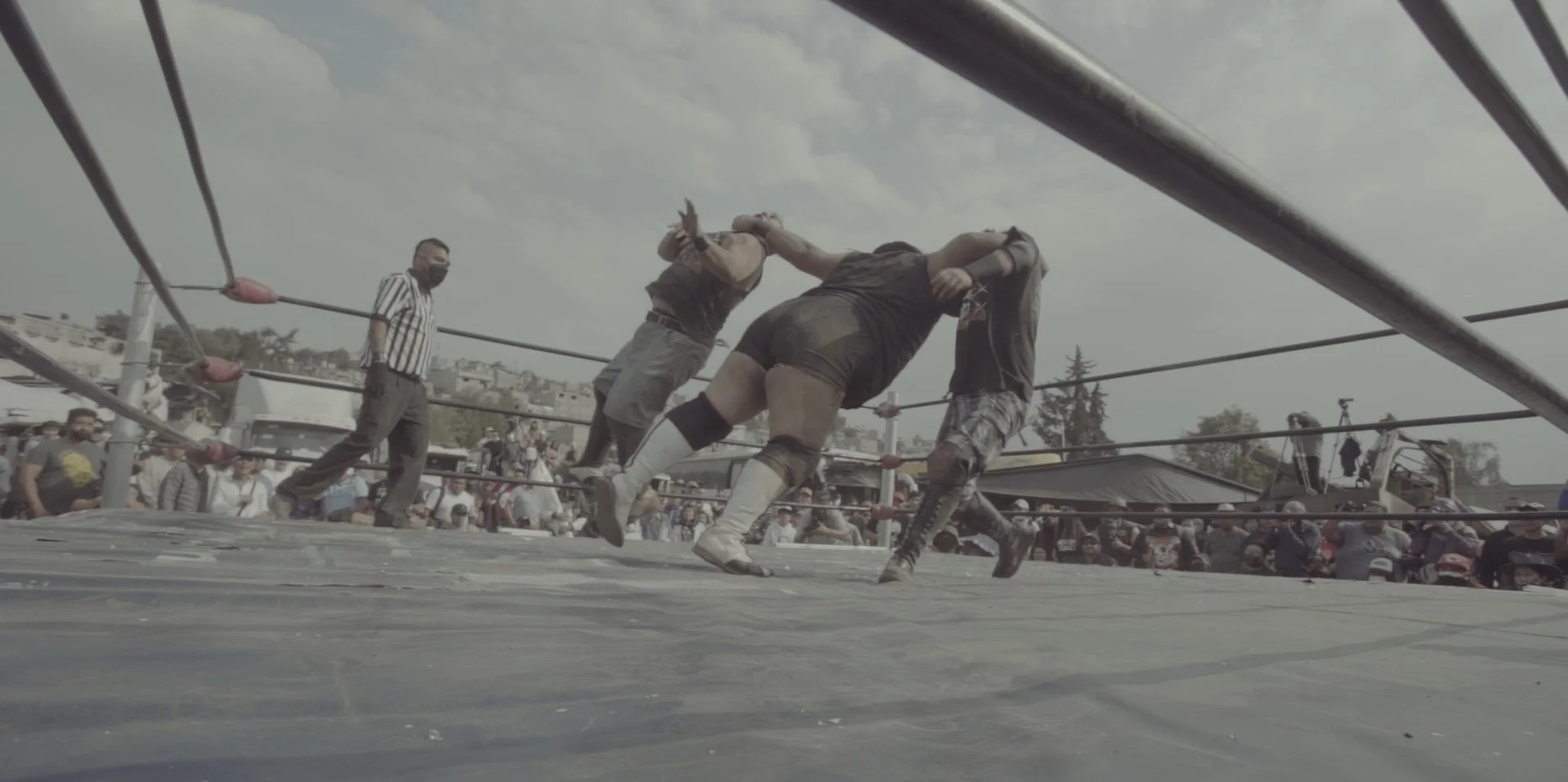 A wrestling match taking place outdoors with two wrestlers engaged, one in black shorts and the other in gray shorts, among spectators and a referee on a wrestling ring.