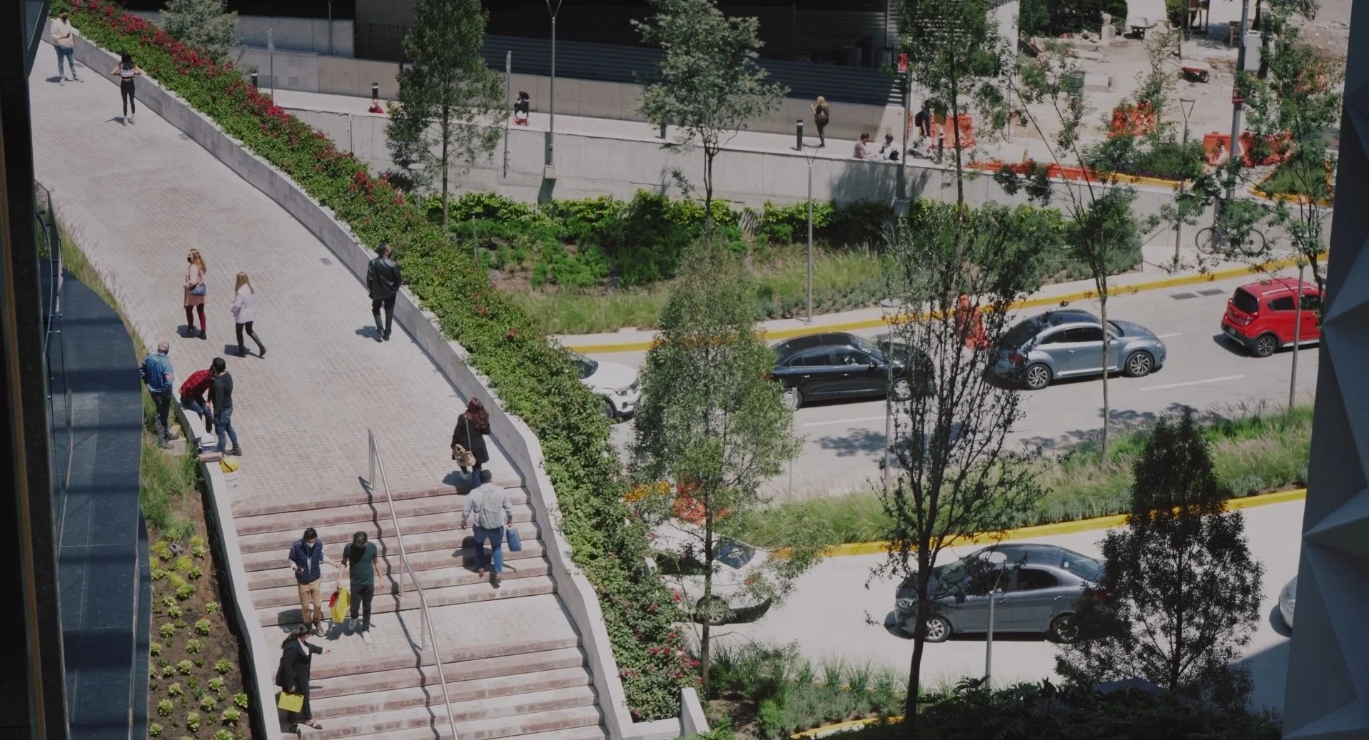 A view of a busy outdoor urban scene with people walking up and down stairs and along pathways, parked cars, trees, and green landscaping.
