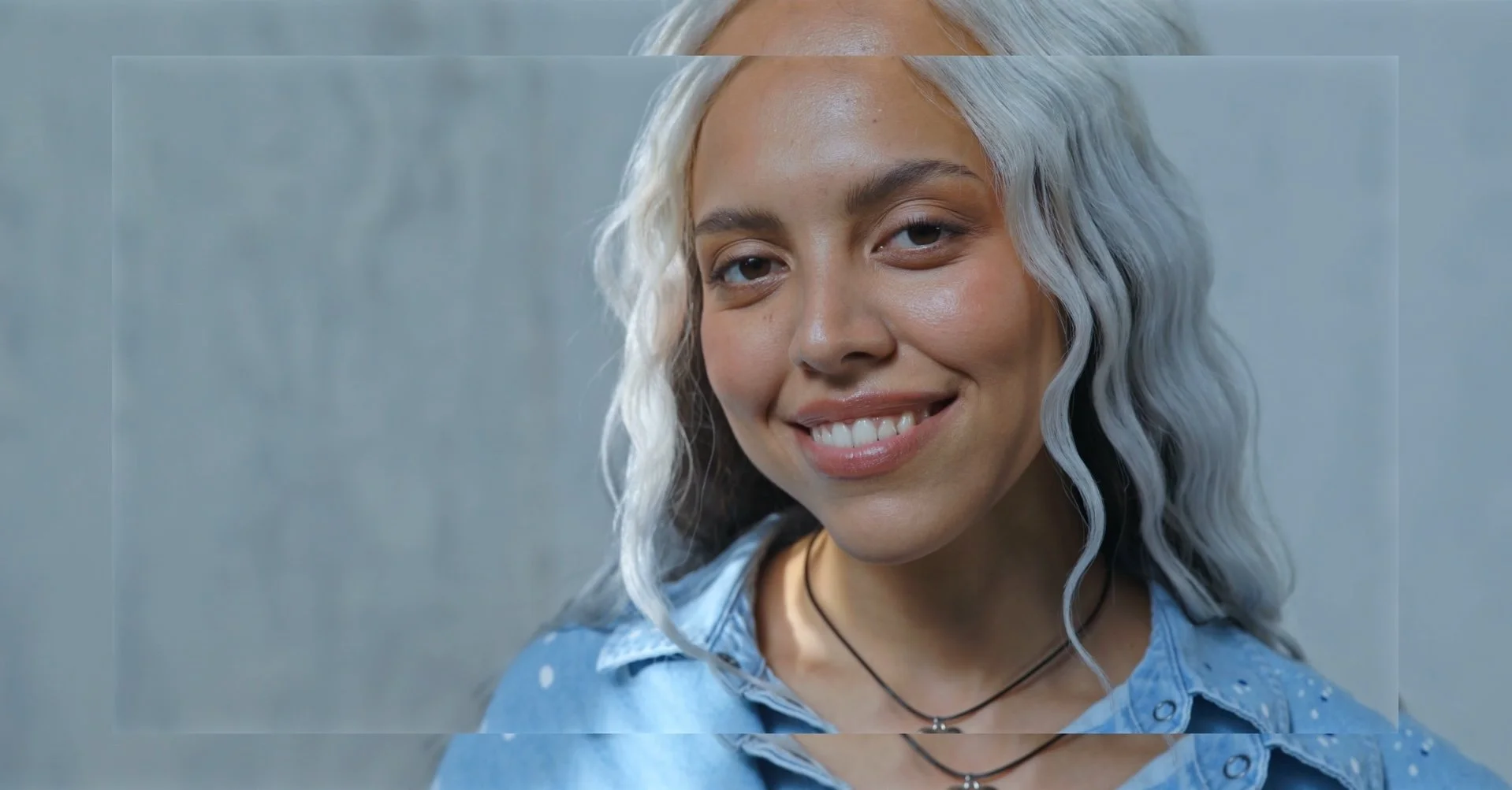A young woman with curly silver hair, smiling, wearing a denim shirt and layered necklaces, standing in front of a gray wall.
