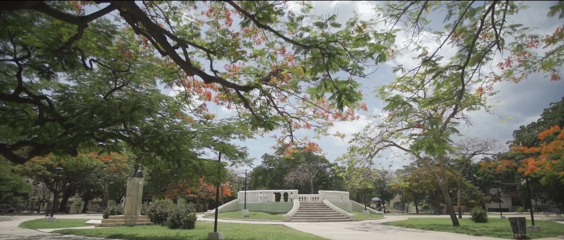 Public park with a white monument, trees with green and orange leaves, cloudy sky, and pathways.