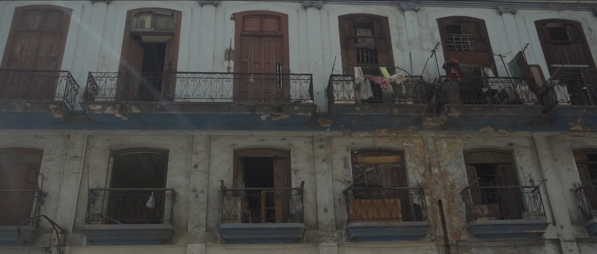 Three-story old building with worn white walls, brown wooden shutters on the windows, and black wrought-iron balconies with laundry hanging to dry. The building appears weathered and in disrepair.