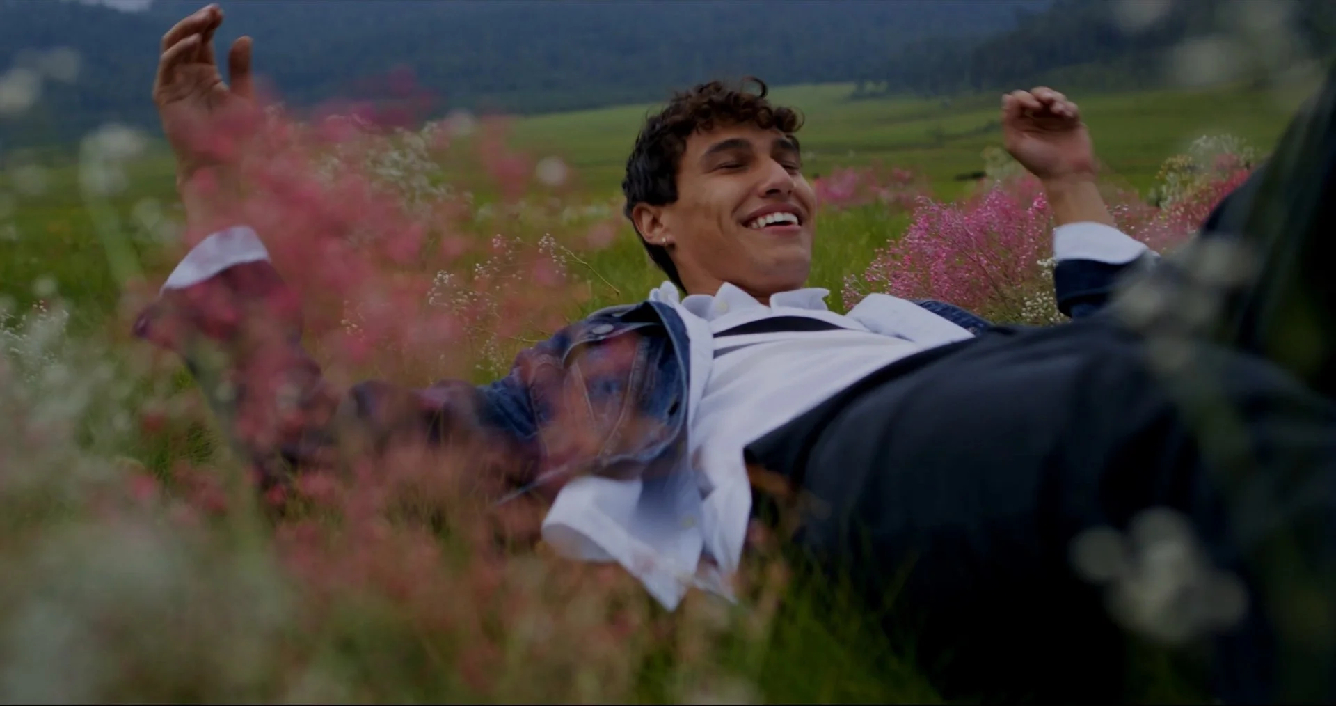 A young man lying in a field of pink and white flowers, smiling with arms raised, during daytime.