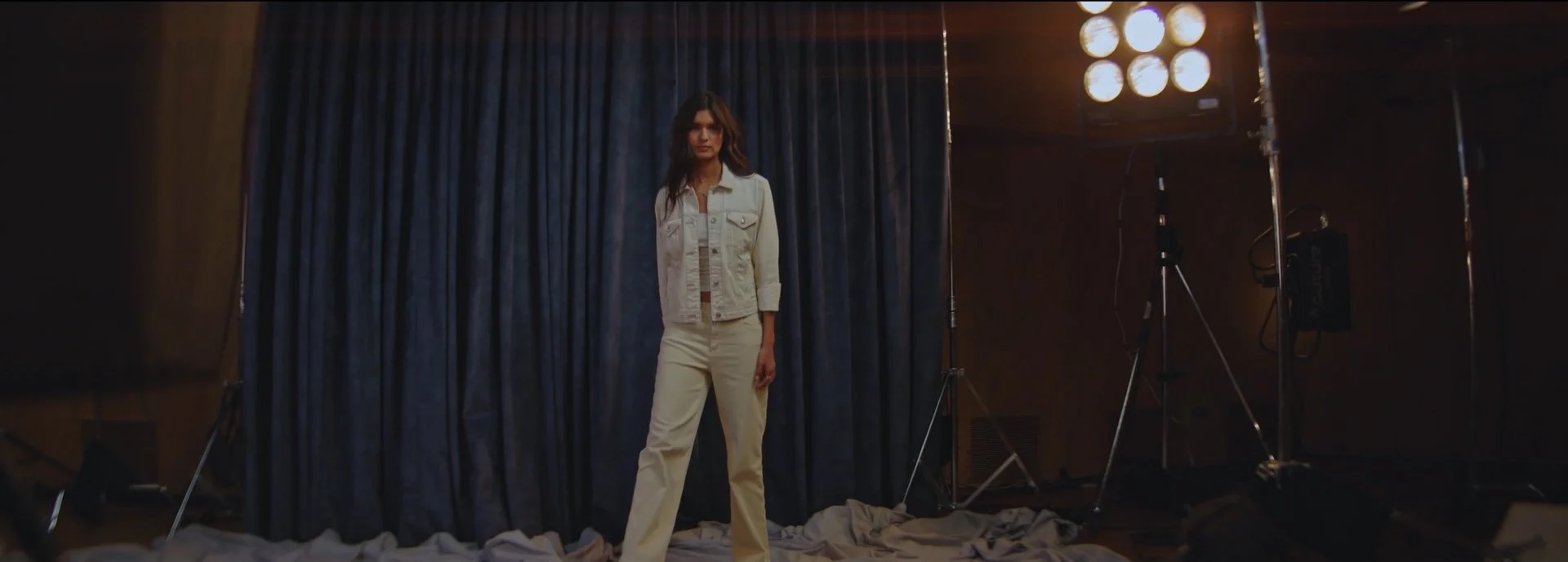 A woman stands on a photography set with a dark curtain backdrop, studio lights to her right, and scattered fabric on the floor.