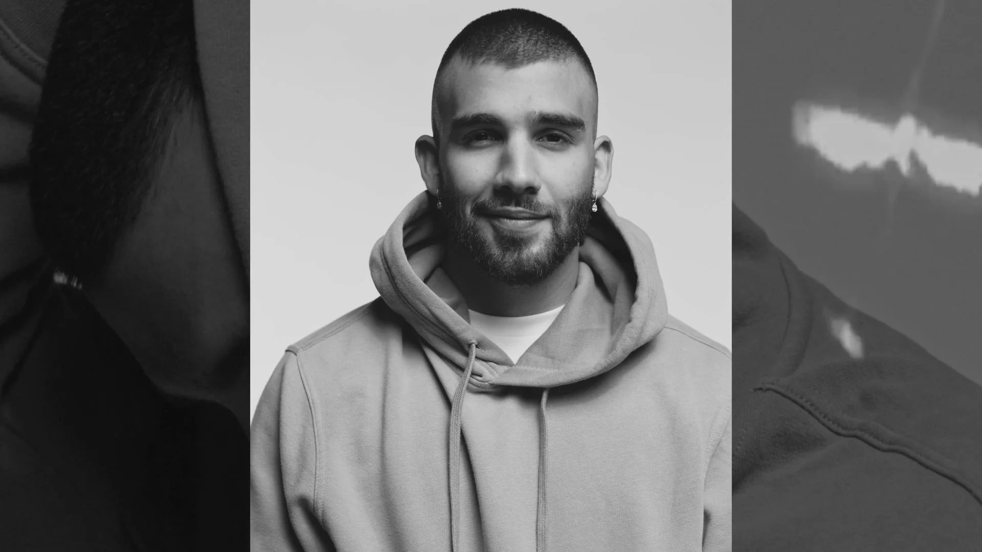Black and white portrait of a man with a beard, short hair, earrings, wearing a hoodie, smiling slightly.