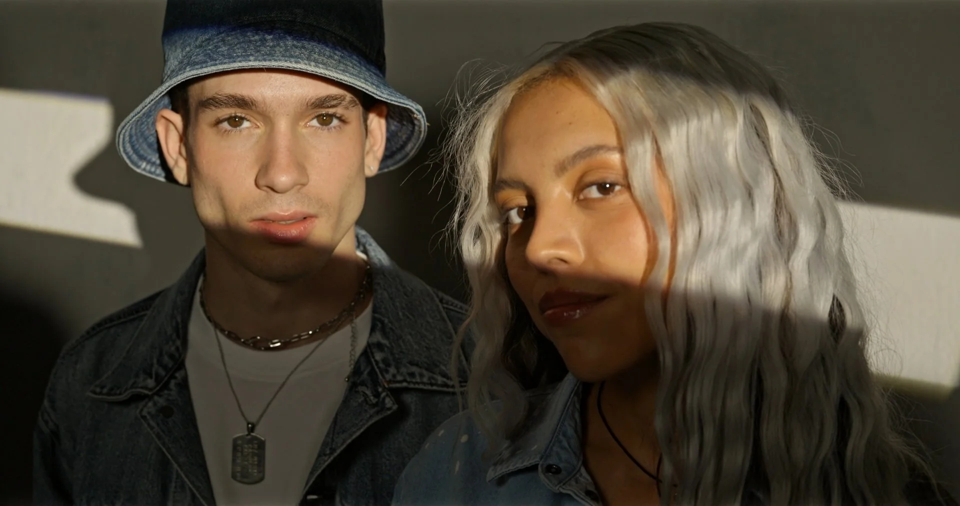 Close-up of a young man and woman standing indoors with shadows cast across their faces, both looking at the camera.