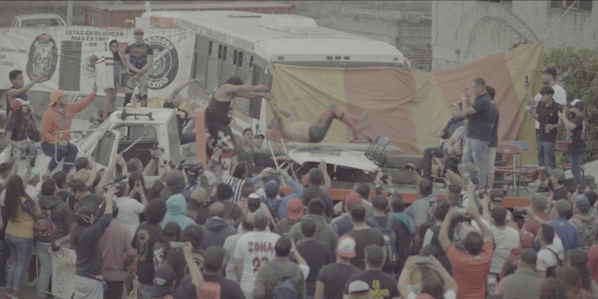 Crowd gathered around a staged event with a wrestling or martial arts stunt, where a shirtless performer is seen mid-air after jumping off a vehicle, as spectators film and cheer.