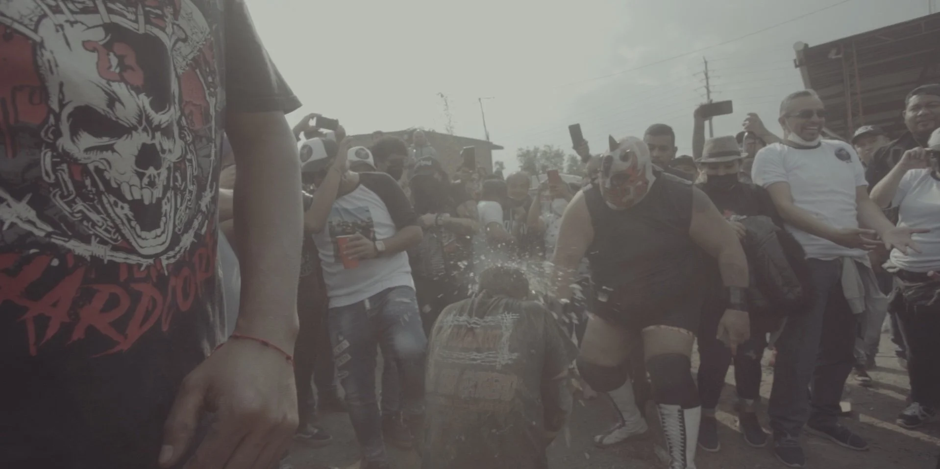 People gathered outdoors during a daytime event, with one person wearing a mask and wrestling gear, and others taking photos or watching.
